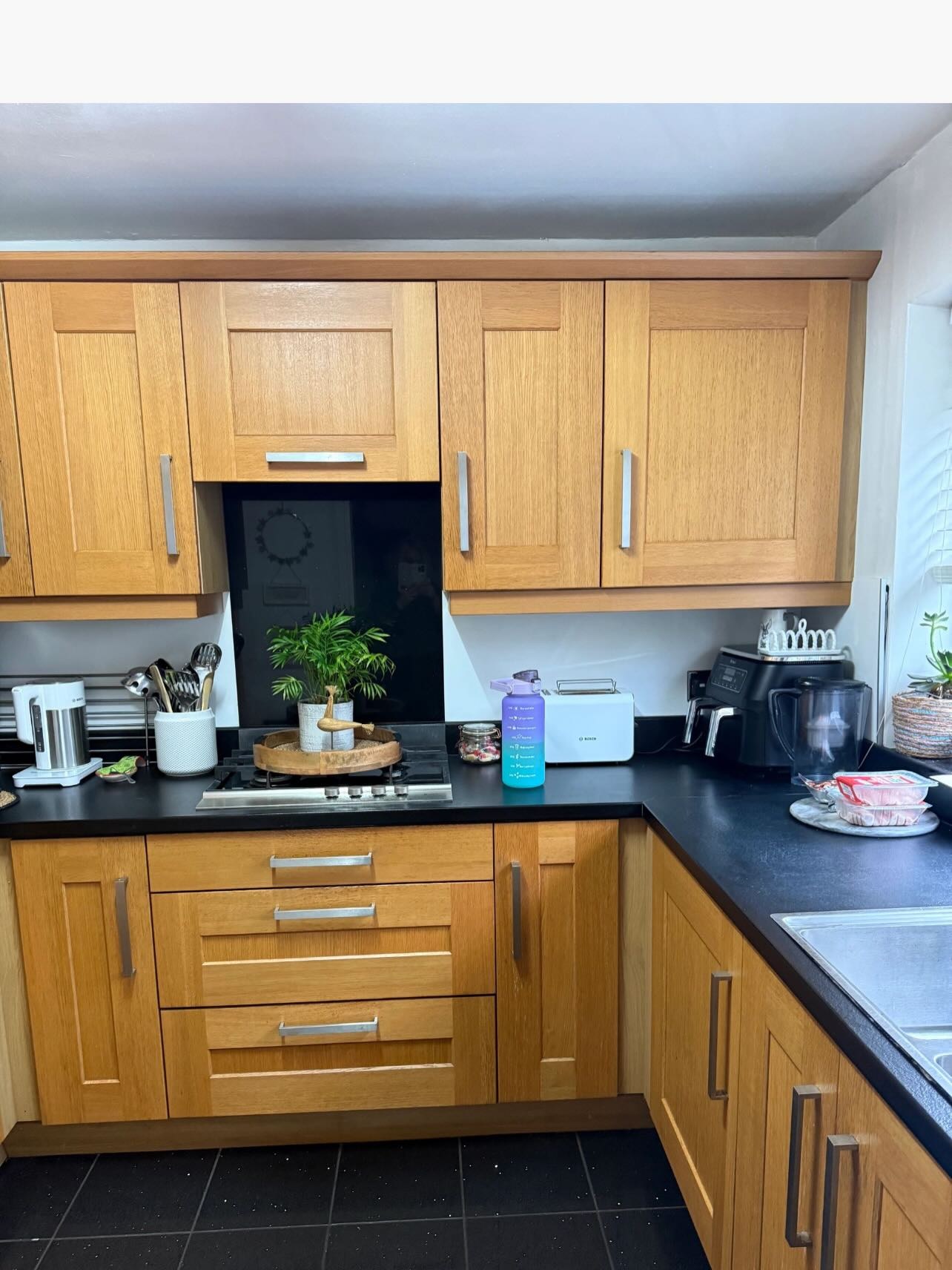 Honey oak shaker kitchen before painting, with a stainless steel hob set into a black laminate counter, black floor tile, and oak upper and lower cabinets catching warm orange light.