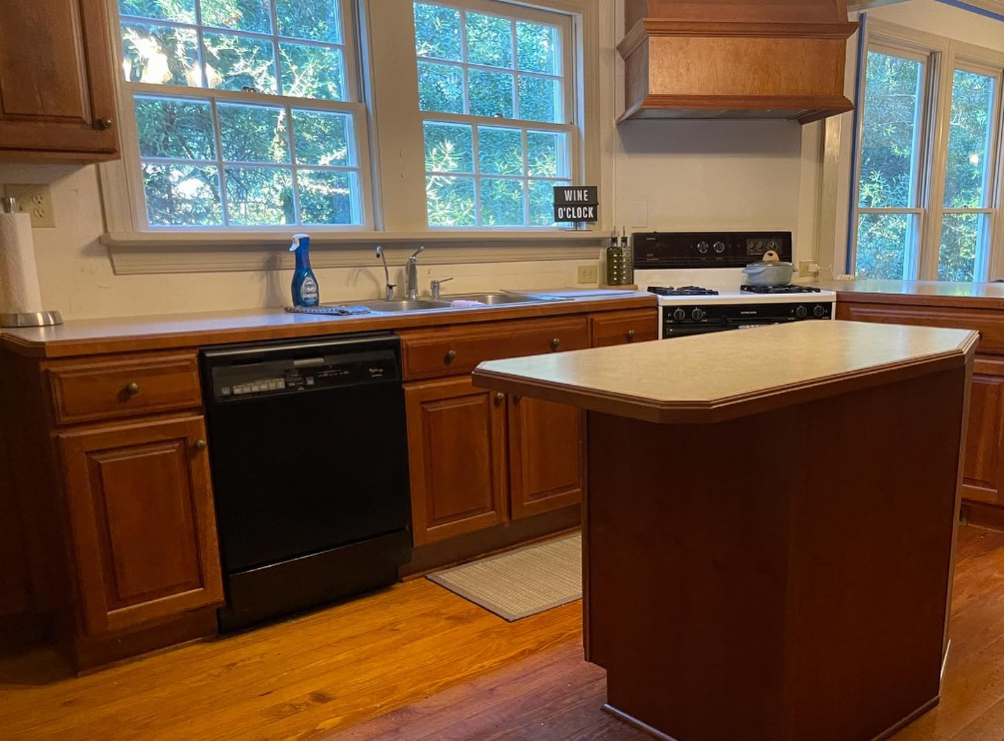 Wide view of a dated kitchen with honey oak cabinets, formica island with angled corners, double stainless sink below two double-hung windows, black Whirlpool dishwasher, almond gas range, wood floors, and a small "wine o'clock" sign on the windowsill.