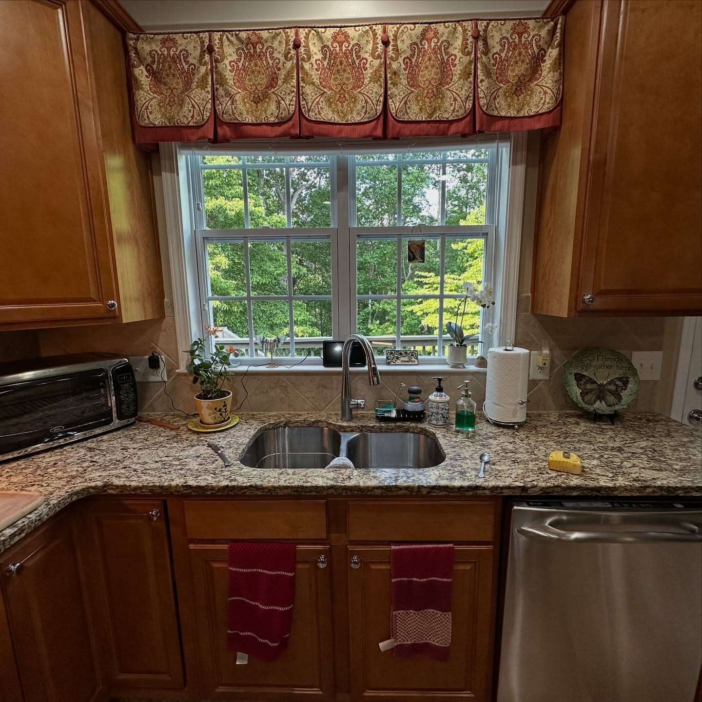 Kitchen sink area with orange oak cabinets, a cream and brown speckled granite counter, diamond-set tile backsplash, undermount stainless sink, and a heavy red and gold paisley valance over the window.