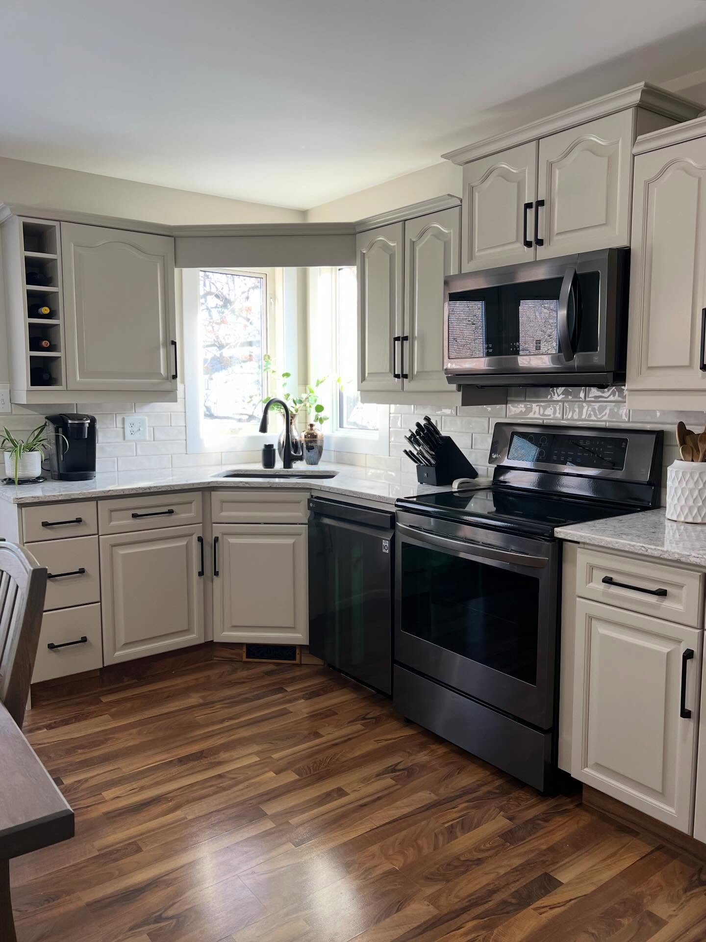 Corner sink in the original kitchen with dark honey oak cabinets, a Keurig on the left counter, speckled brown laminate countertop, chrome faucet, and a bay window looking out at bare trees.