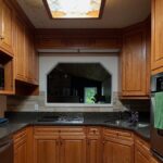 Wide view of the honey oak U-shaped kitchen before renovation, with cathedral raised-panel cabinets, a stained glass fluorescent ceiling light box, dark speckled granite counters, a built-in stainless wall oven on the right, a cooktop and corner sink along the back wall, warm tan stone-look tile floor, and a pass-through window above the counter looking into a stone fireplace living room.