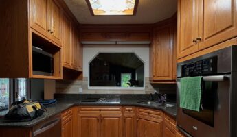 Wide view of the honey oak U-shaped kitchen before renovation, with cathedral raised-panel cabinets, a stained glass fluorescent ceiling light box, dark speckled granite counters, a built-in stainless wall oven on the right, a cooktop and corner sink along the back wall, warm tan stone-look tile floor, and a pass-through window above the counter looking into a stone fireplace living room.