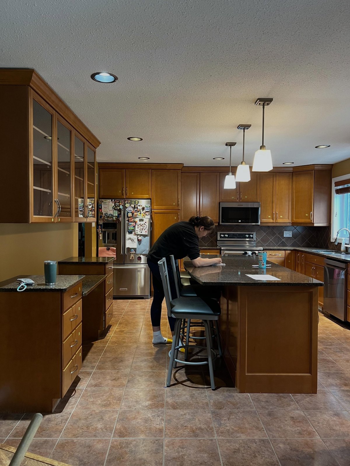 Wide view of a kitchen with orange honey oak raised-panel cabinets, dark speckled granite counters, brown diamond-pattern tile backsplash and warm terracotta floor tile, with a woman wiping down the island before the makeover begins.