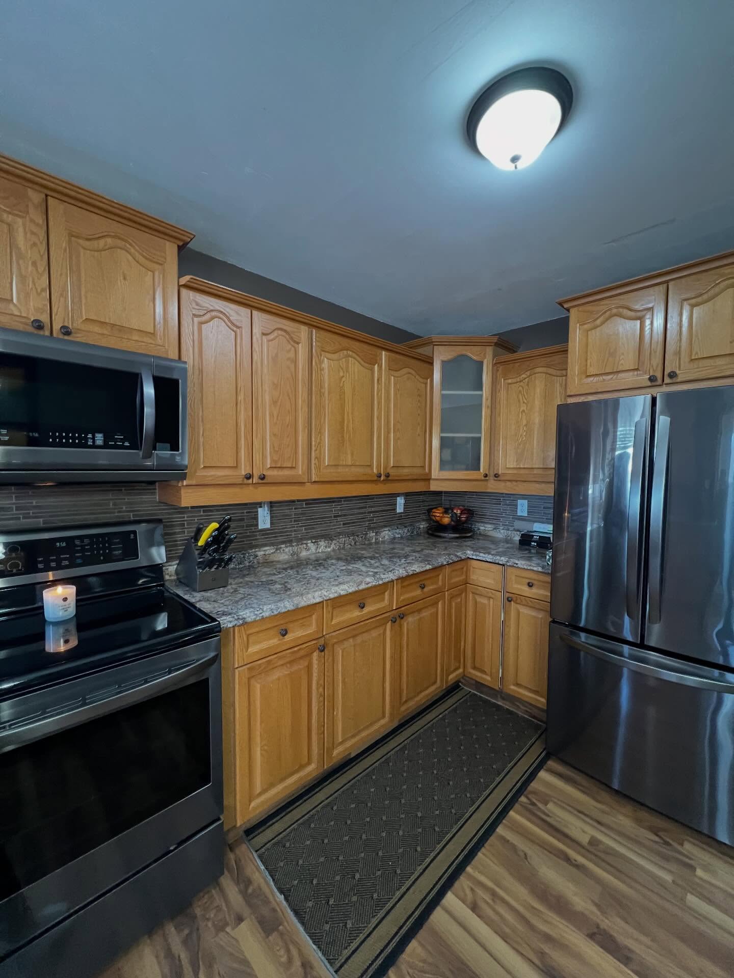 Wide view of a 90s kitchen with dark honey oak cabinets, arched raised-panel cabinet doors, speckled brown granite counters, a dark linear tile backsplash, and gray walls that pull the whole room down.