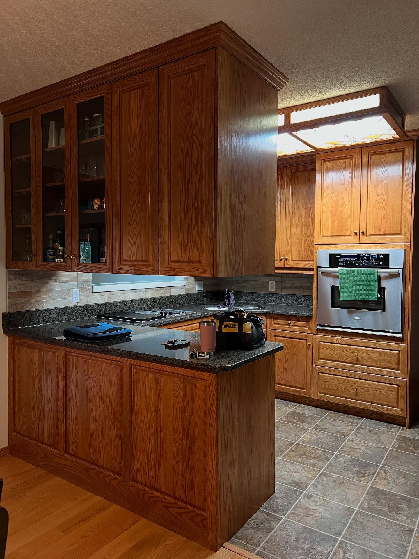 Honey oak kitchen peninsula with cathedral raised-panel bar front, glass-front upper cabinets holding glassware and bottles above the back counter, dark speckled granite, a built-in stainless wall oven on the right, and a fluorescent ceiling light box above, with a DeWalt tool bag on the counter.