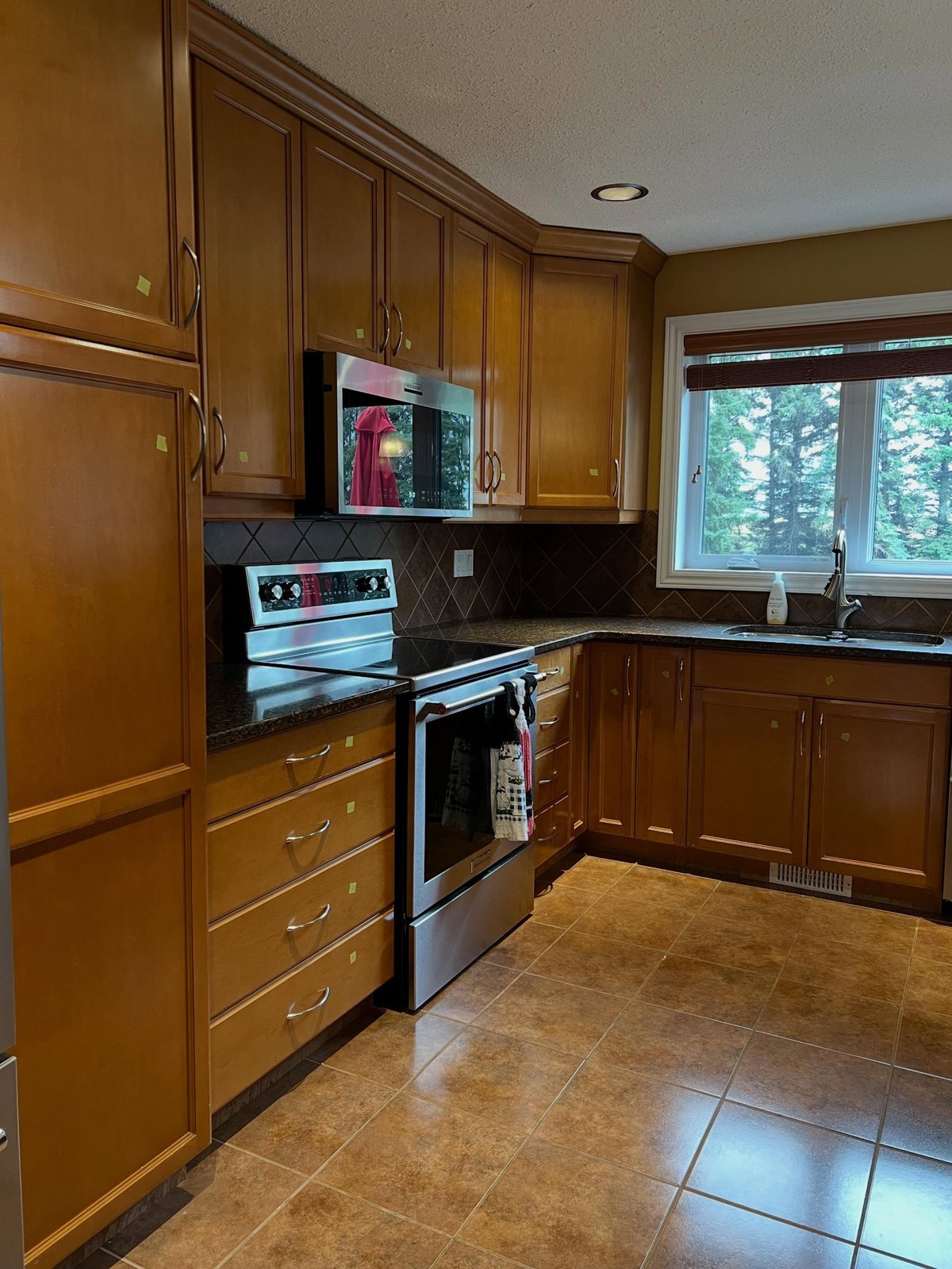 Close-up of the kitchen wall holding the electric stove, microwave and window over the sink, lined in orange honey oak cabinets with brown diamond backsplash tile and dark speckled granite counters.
