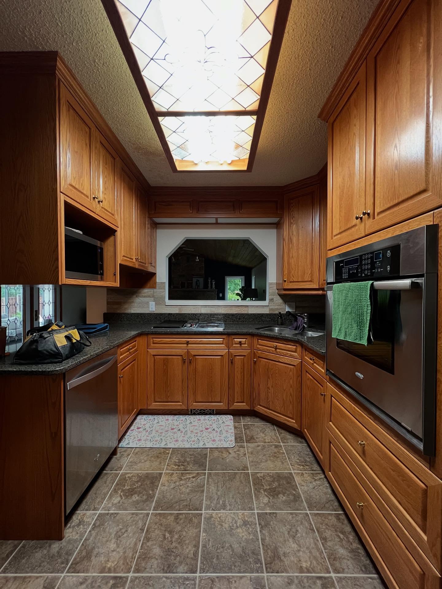 U-shaped honey oak kitchen with cathedral raised-panel doors, dark speckled granite counters, a stained glass fluorescent ceiling light box, tan stone-look tile floor, a built-in wall oven on the right, and a pass-through window to a stone fireplace living room.