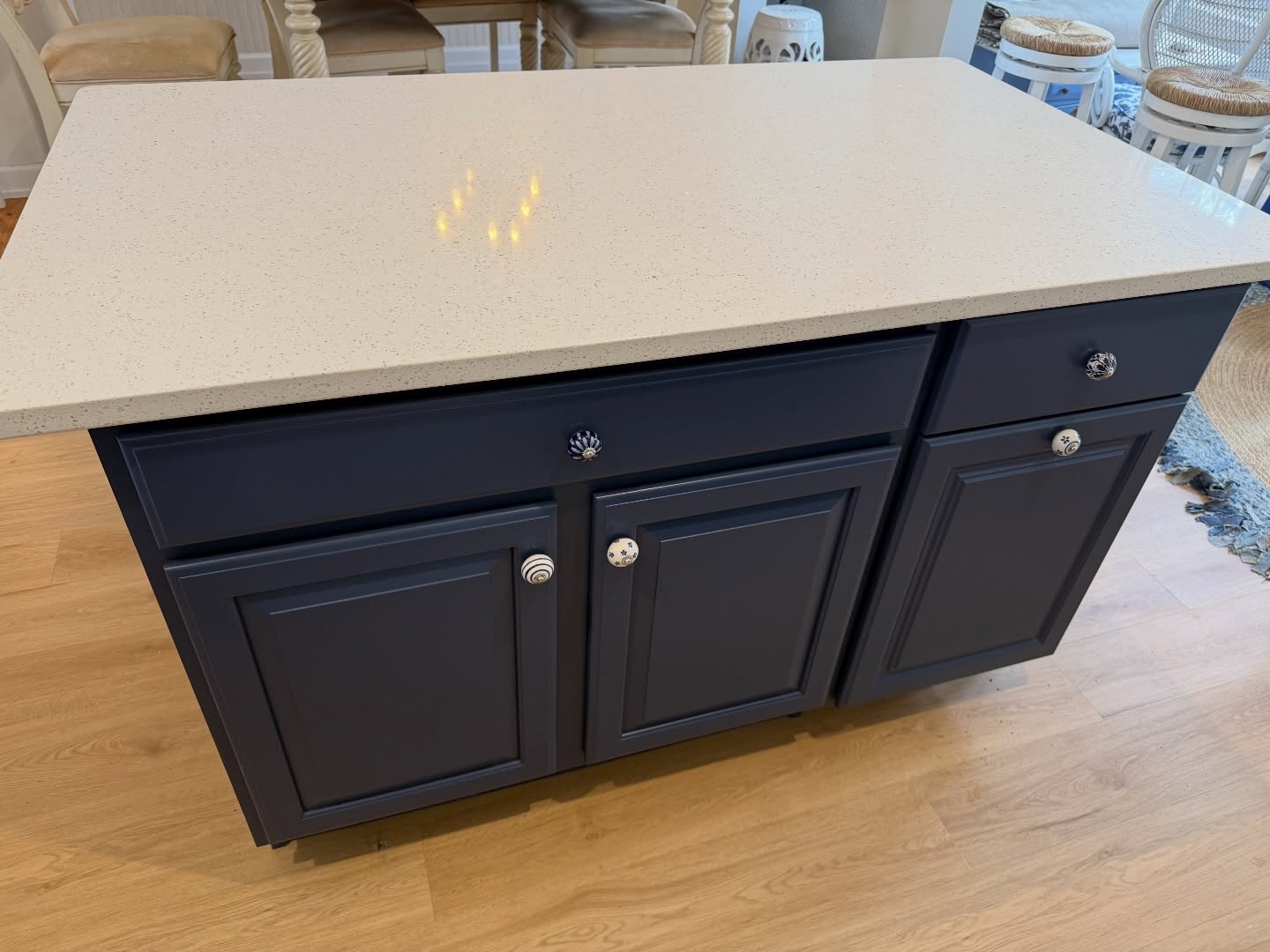 Close view of a kitchen island painted deep navy blue in Sherwin-Williams Indigo Batik, with a white quartz speckled top, decorative ceramic knobs, and light oak flooring.