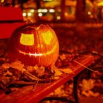 A carved jack-o-lantern with triangle eyes and a jagged smile glows warm orange from inside, sitting on a dark red wooden park bench piled with dry fallen autumn leaves, with soft blurred city lights behind it at night.