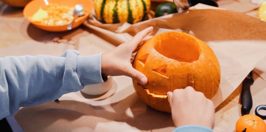 Child in a light blue sweatshirt carving a jaw into a jack-o-lantern pumpkin on a kraft paper covered table, surrounded by scooped pumpkin pulp in an orange bowl, a small decorative gourd with parsley on top, a carving knife, scissors, and a tiny clementine drawn with a jack-o-lantern face.