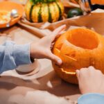 Child in a light blue sweatshirt carving a jaw into a jack-o-lantern pumpkin on a kraft paper covered table, surrounded by scooped pumpkin pulp in an orange bowl, a small decorative gourd with parsley on top, a carving knife, scissors, and a tiny clementine drawn with a jack-o-lantern face.