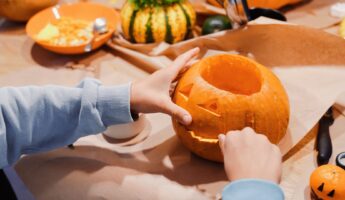 Child in a light blue sweatshirt carving a jaw into a jack-o-lantern pumpkin on a kraft paper covered table, surrounded by scooped pumpkin pulp in an orange bowl, a small decorative gourd with parsley on top, a carving knife, scissors, and a tiny clementine drawn with a jack-o-lantern face.