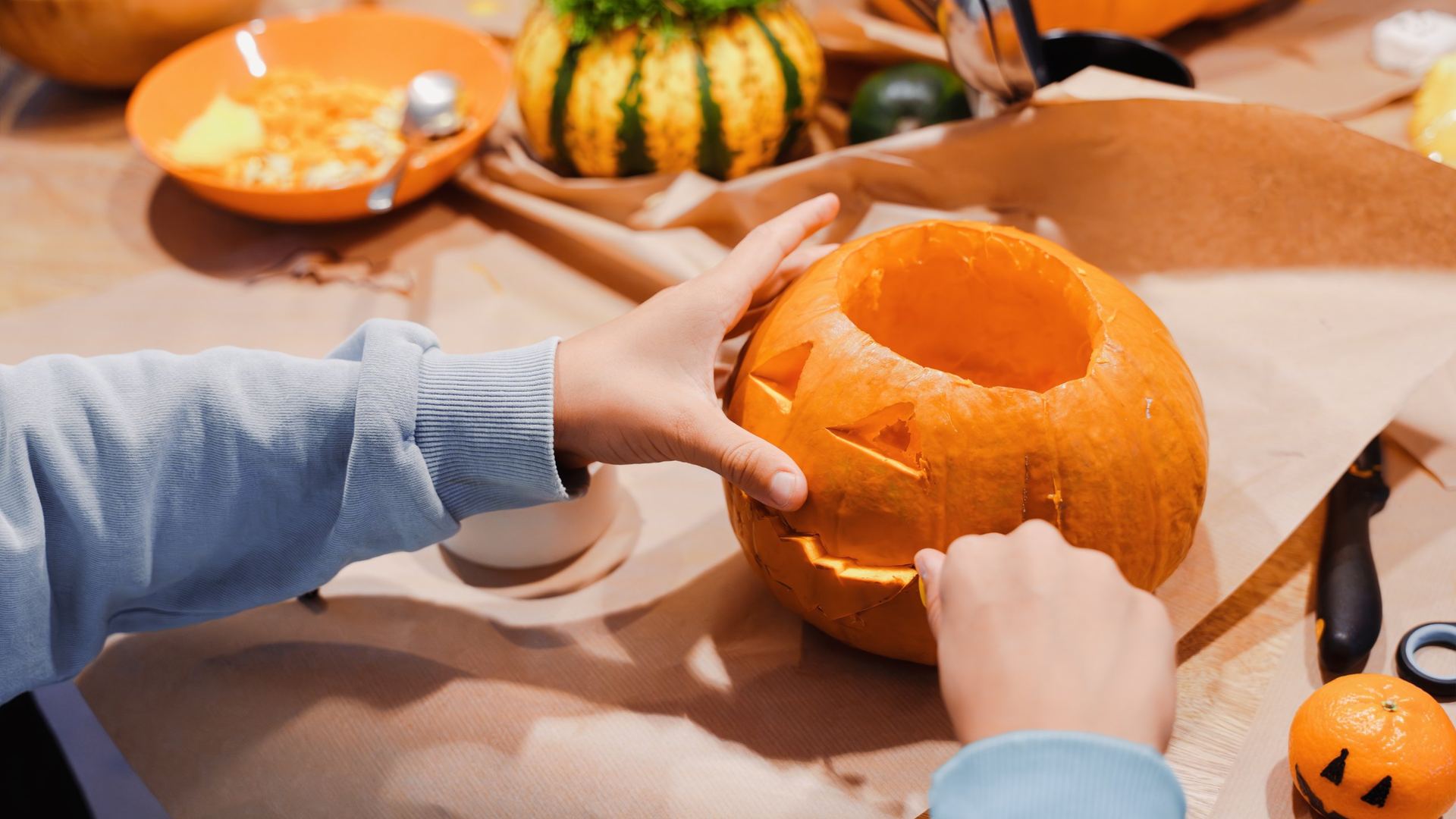 Child in a light blue sweatshirt carving a jaw into a jack-o-lantern pumpkin on a kraft paper covered table, surrounded by scooped pumpkin pulp in an orange bowl, a small decorative gourd with parsley on top, a carving knife, scissors, and a tiny clementine drawn with a jack-o-lantern face.