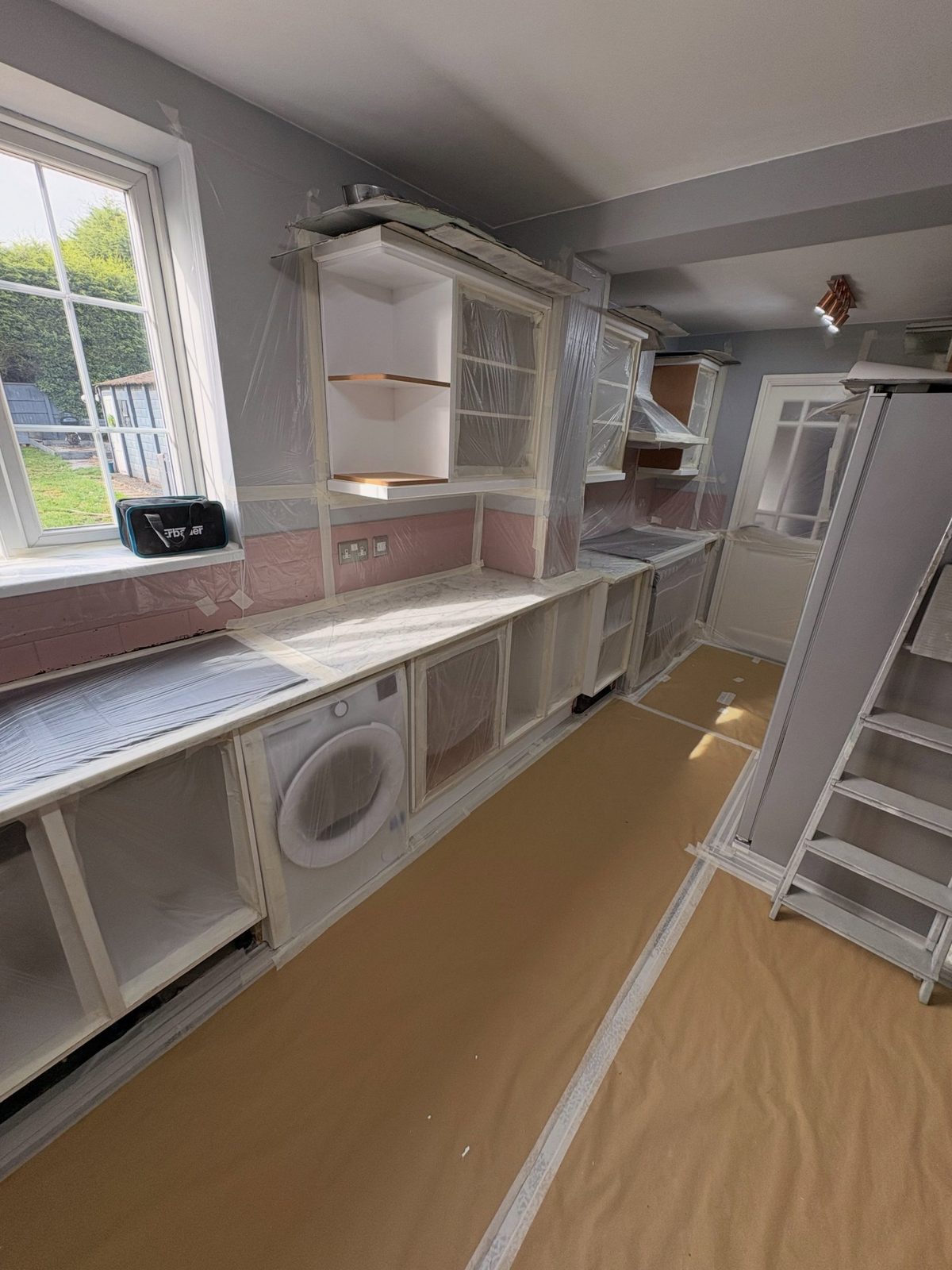 Kitchen cabinets covered in plastic sheeting and masking tape in preparation for professional spray painting, with brown floor paper laid down and the marble counter protected.