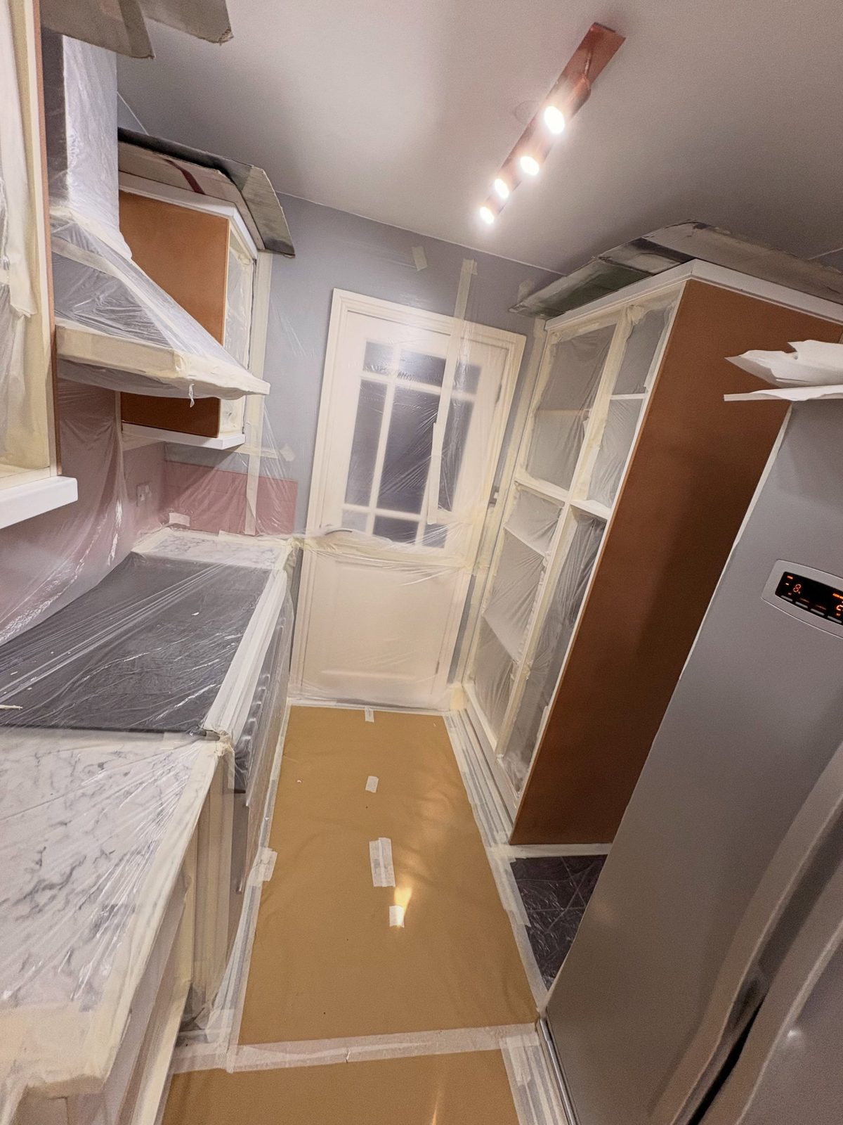 Cabinet shells and doors showing an orange-toned primer coat with the fridge and ceiling light visible, floor still papered and plastic sheeting over surfaces.