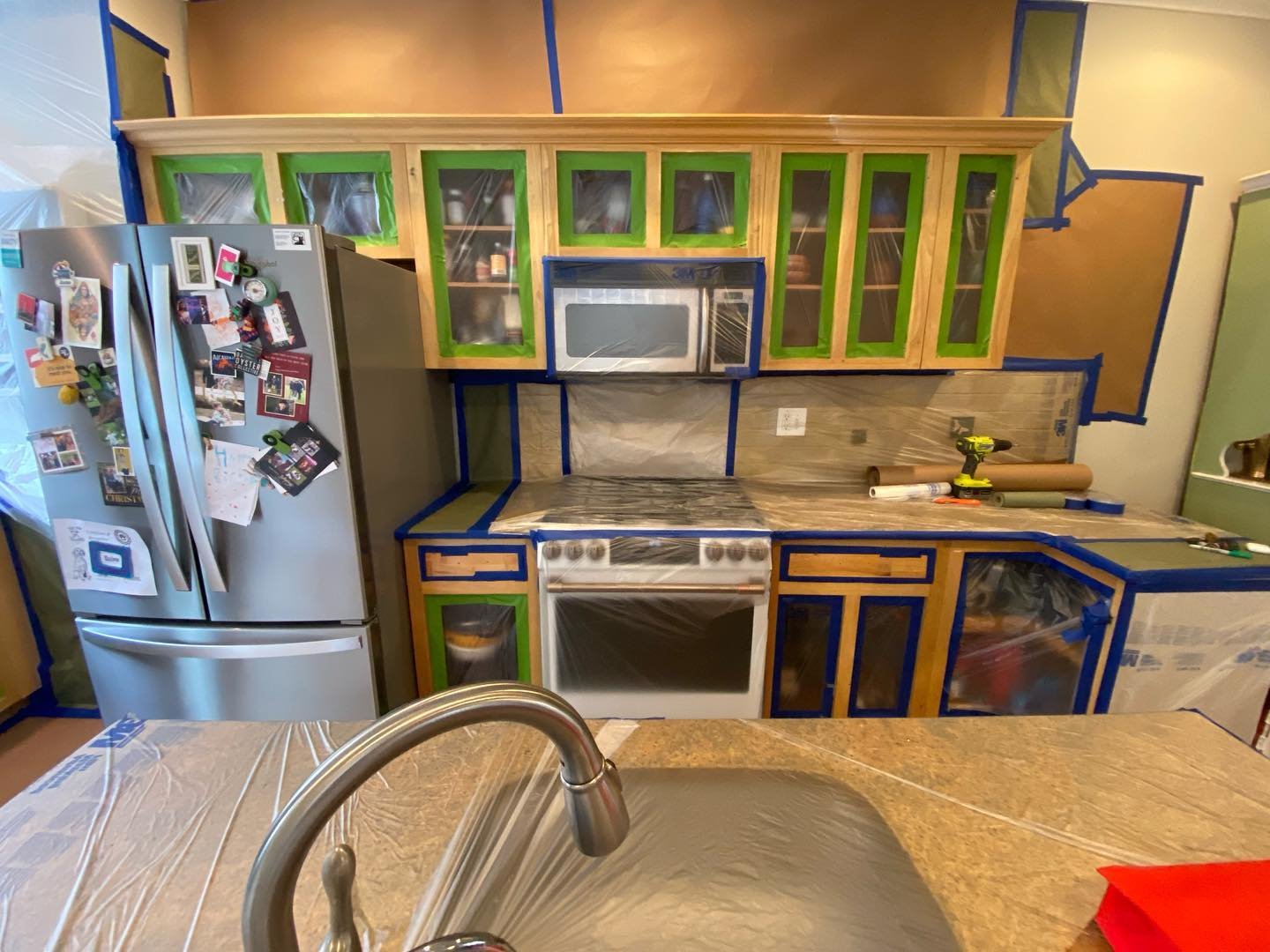 Kitchen in mid-renovation with every cabinet door removed, interior cabinet frames taped off with green painters tape and plastic sheeting, the fridge and range covered in masking paper.