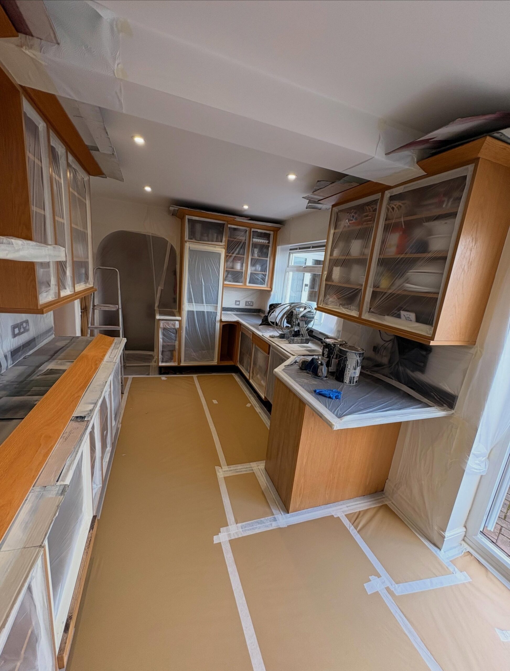 Oak kitchen fully masked with brown floor paper and plastic sheeting over cabinets, worktops, and ceiling, with spray gear on the counter mid prep.