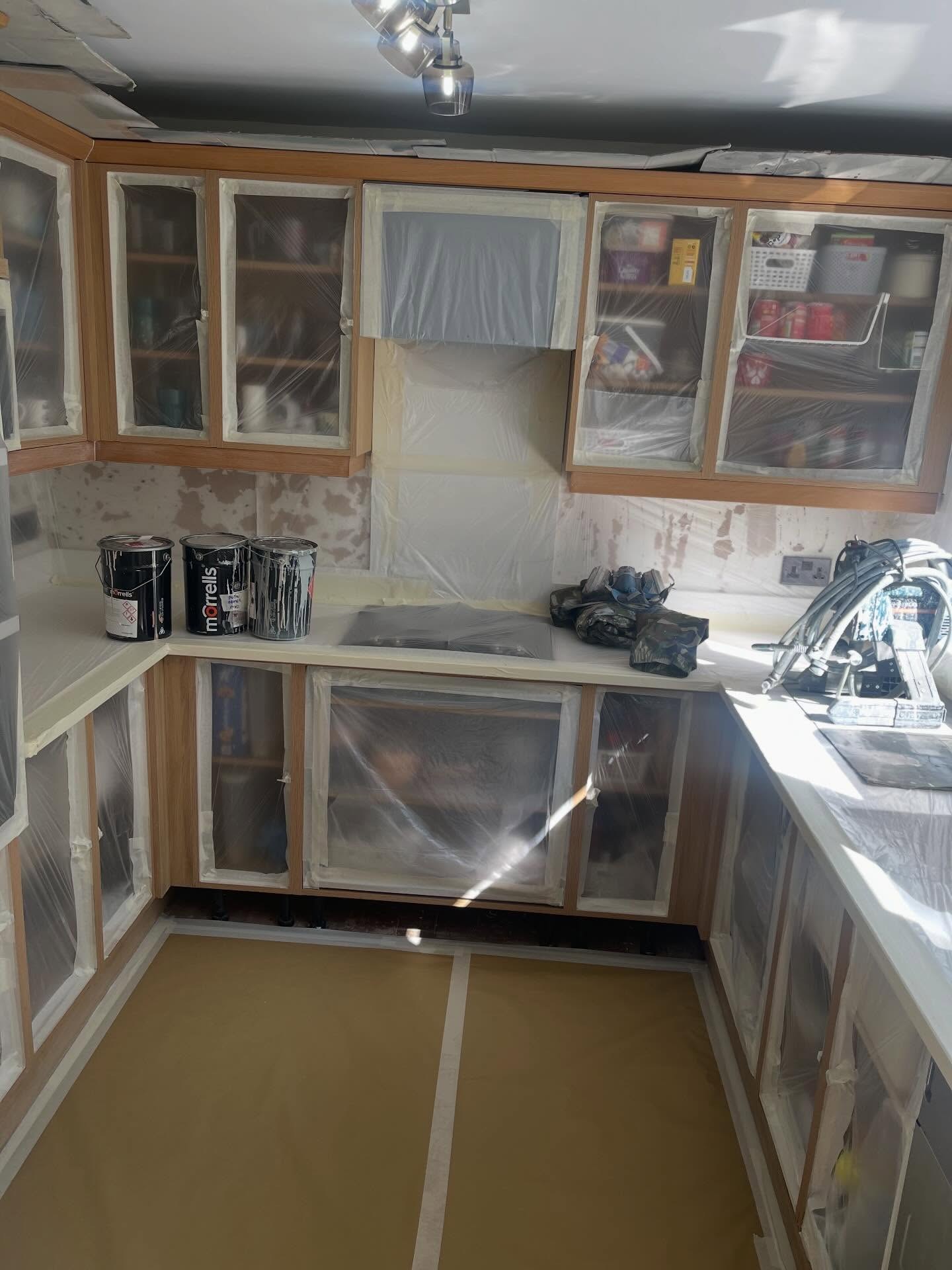 Three black Morrells paint tins sitting on a masked white worktop with the oak cabinets fully wrapped in plastic, mid prep stage before top coats went on.