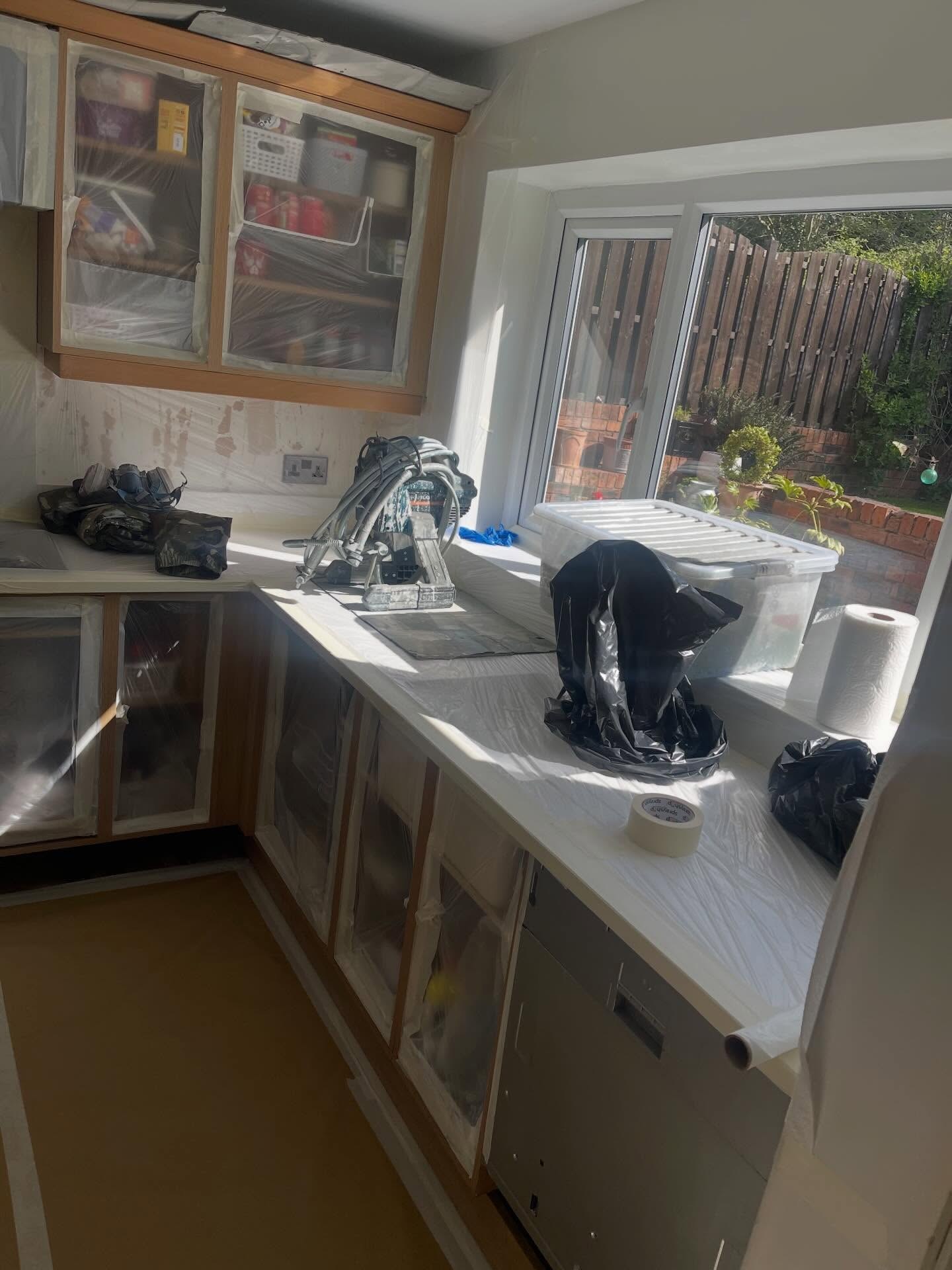 Mid prep kitchen with cabinet doors and worktops wrapped in plastic sheeting and painters tape, an airless sprayer and respirator sitting on the counter near the window, new white worktops already fitted.