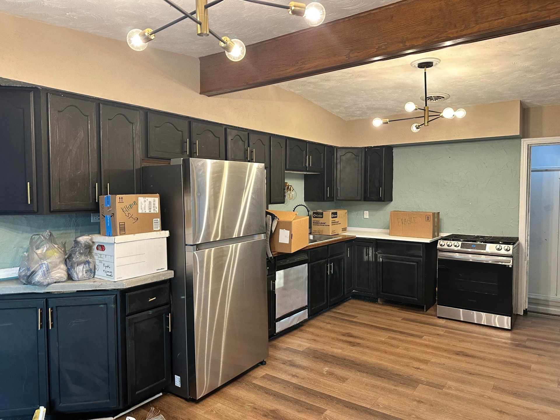 Kitchen during remodel with the new stainless fridge and gas range delivered and boxes stacked on the counters while the old black cabinets, mint stucco walls and the original wood ceiling beam remain in place.