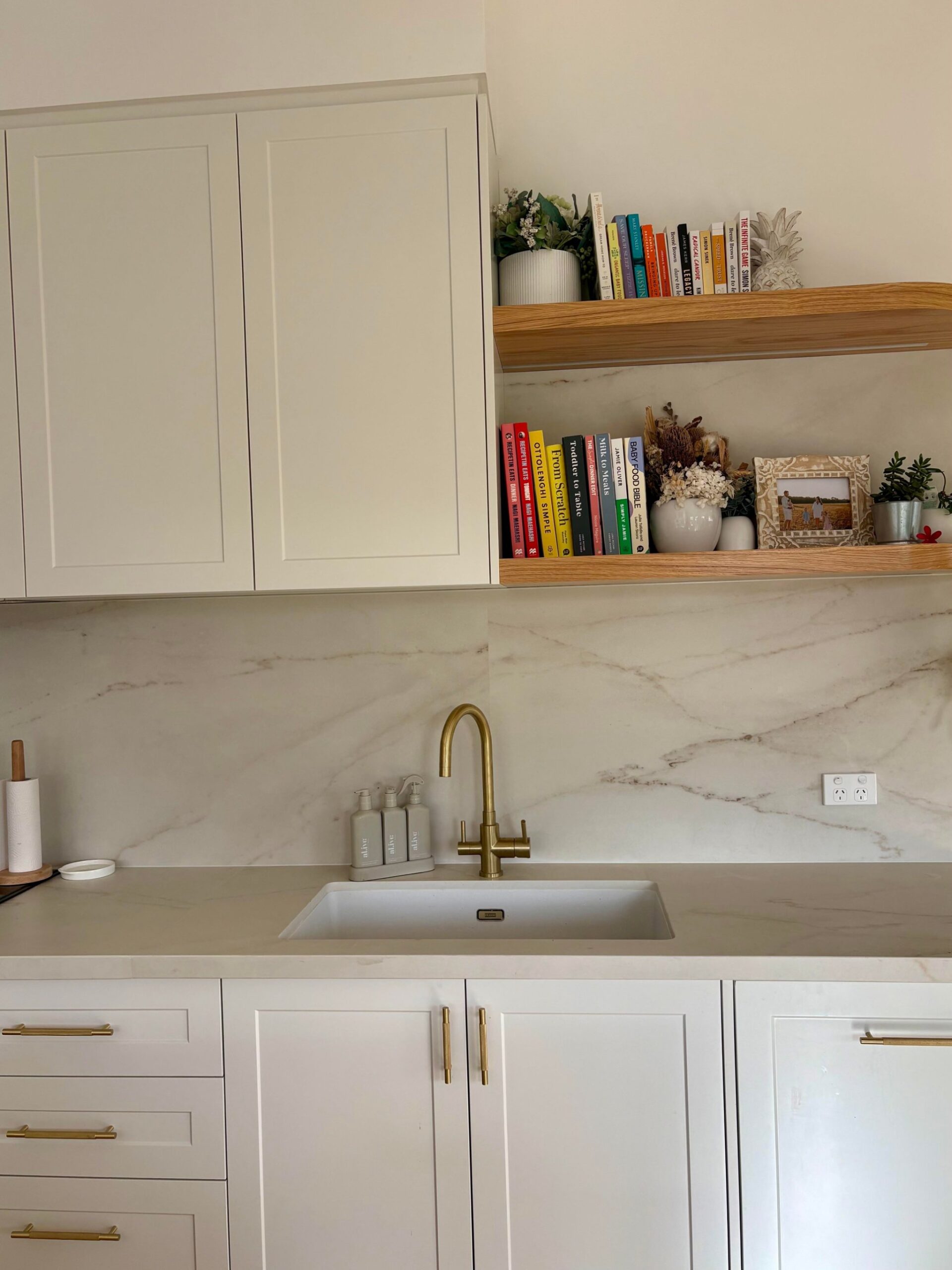 Undermount white sink with a tall brass goose-neck tap, cream Dekton counter, marble backsplash, and two floating oak shelves holding cookbooks and plants.