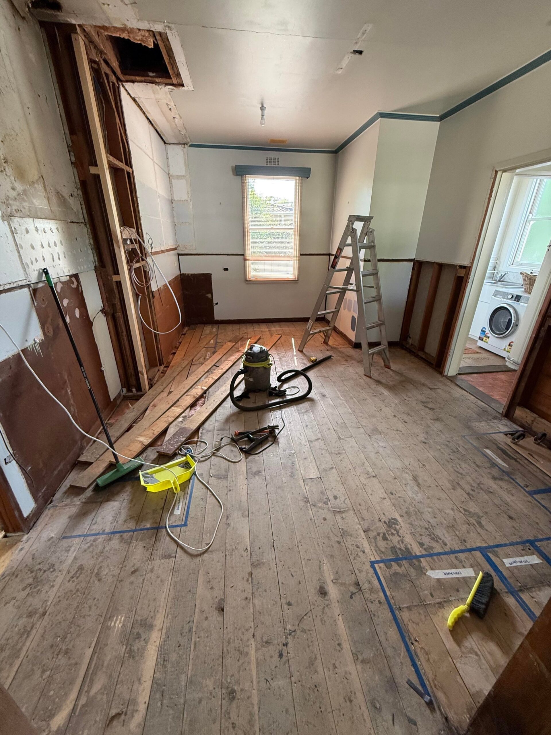 Gutted kitchen with bare wood floorboards, blue tape layout marks, a shop vacuum, a ladder, loose boards, and opened walls showing framing.