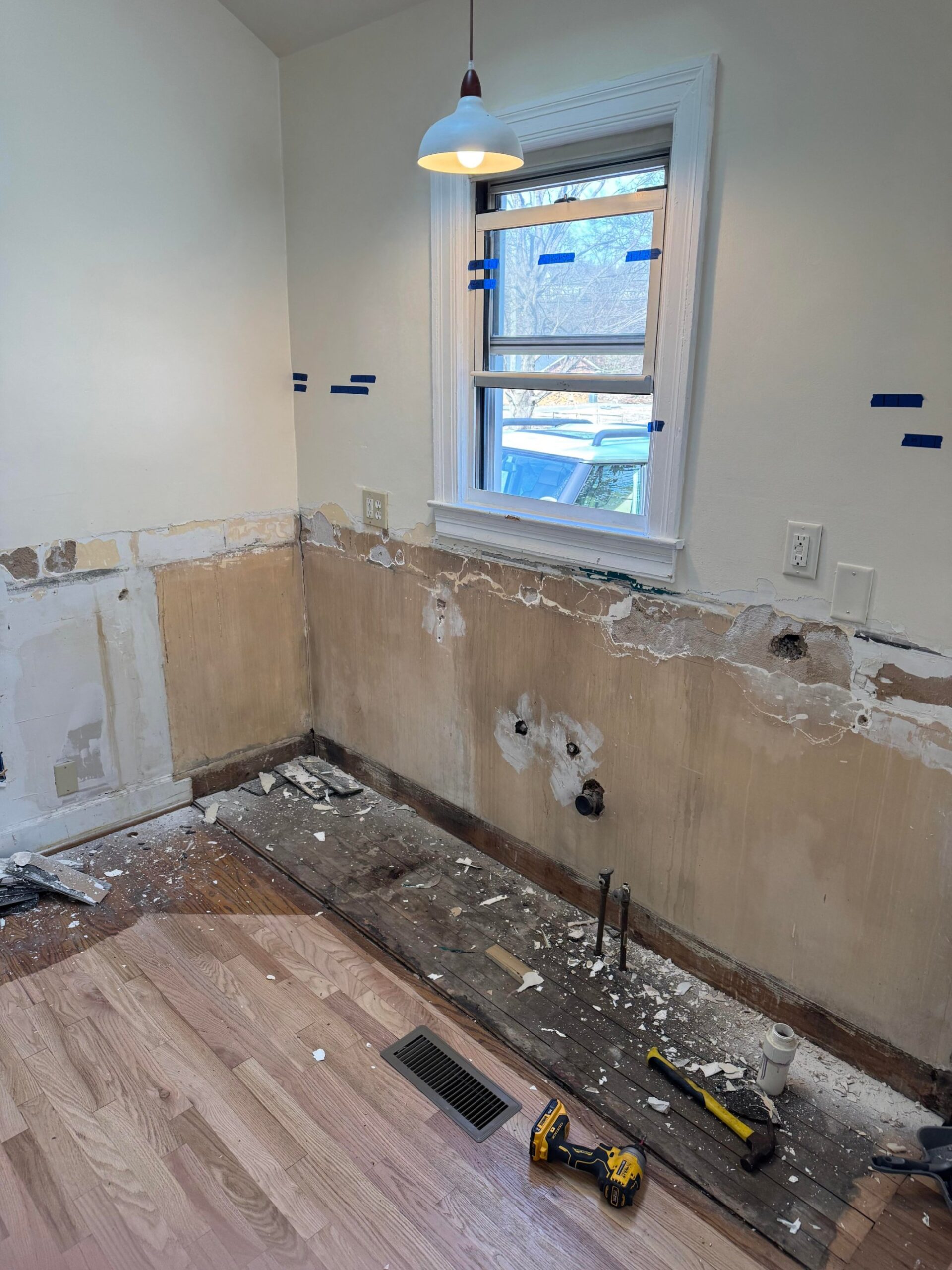 Half-stripped kitchen wall showing bare plaster, exposed water pipes near the floor, old patched holes, and tools on the floor by the window.