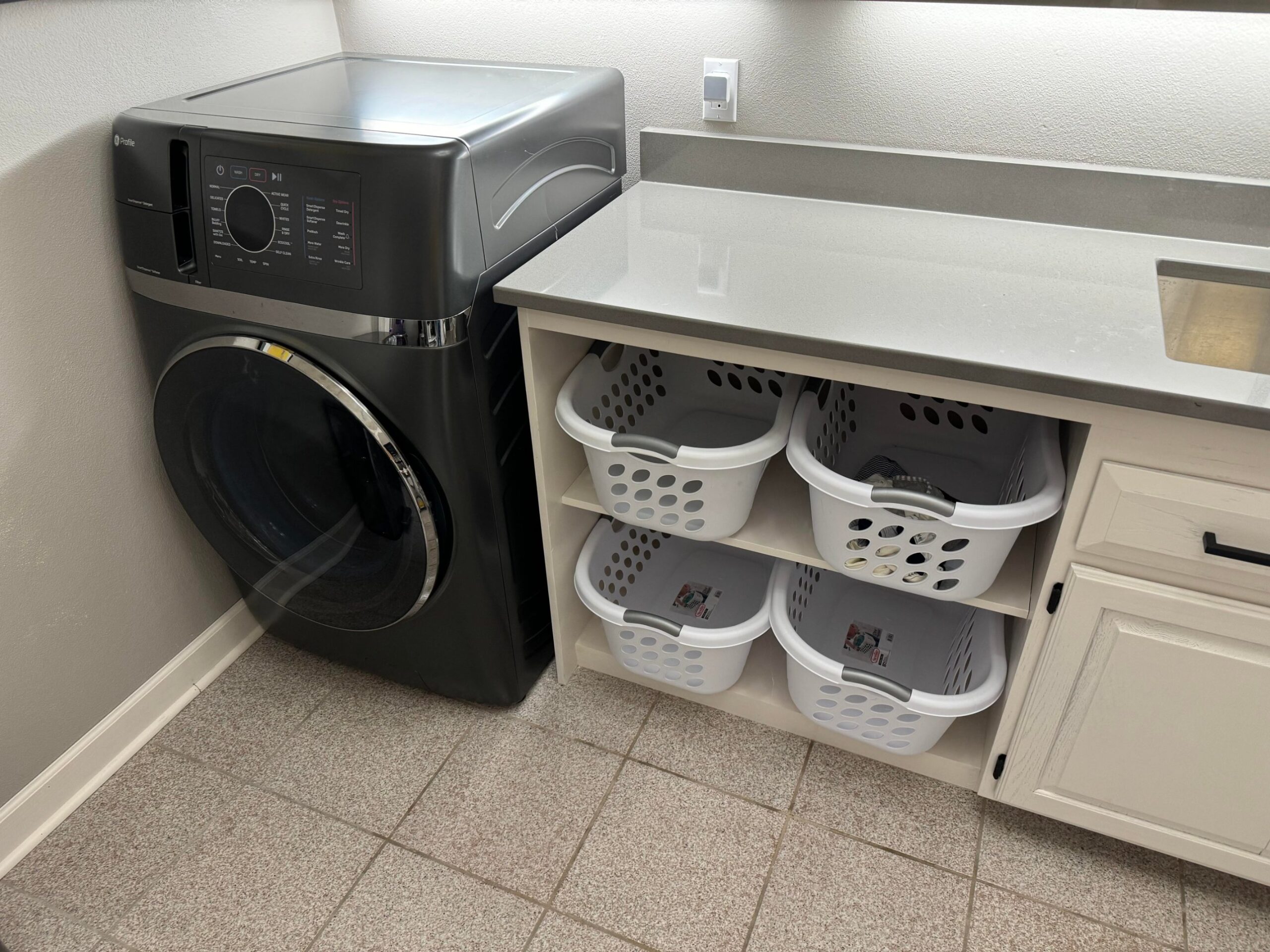 Finished laundry room with black GE Profile all-in-one washer dryer on the left and four white plastic laundry baskets stacked two by two inside built-in cubby shelves, gray quartz counter above, tile floor.