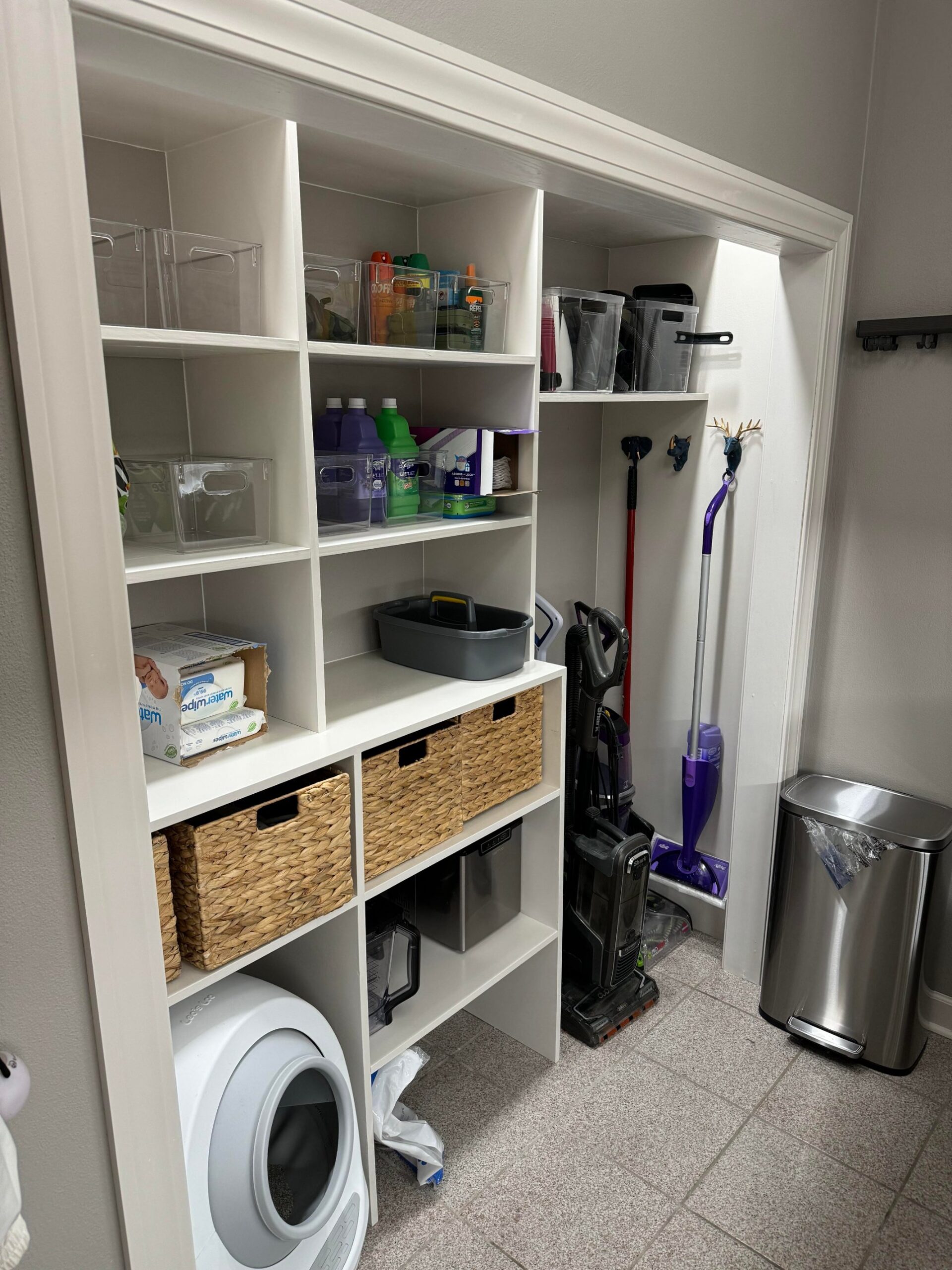 Floor to ceiling white built in closet with cubby shelving on the left holding clear bins with detergents, water wipes, Swiffer refills and insect spray, three woven baskets on a middle shelf, and an upright vacuum, carpet cleaner, broom and Swiffer lined up on the right next to a stainless step trash can.