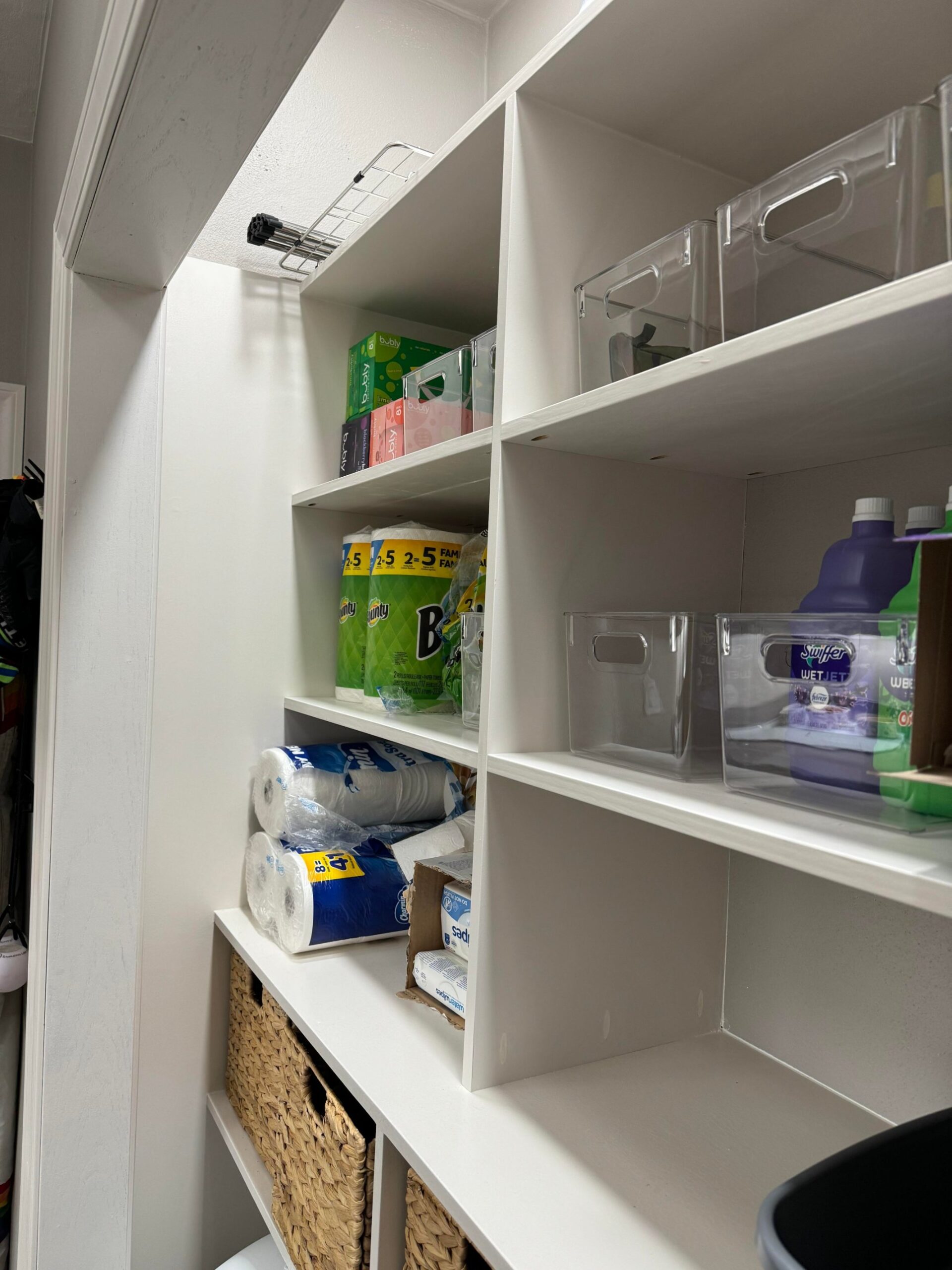 Angled side view of the pantry side of the built in closet showing white plywood cubby shelving stacked four high, loaded with Bubly sparkling water boxes, Bounty paper towel rolls, Charmin toilet paper, Swiffer Wet Jet refills, water wipes and clear organizer bins.