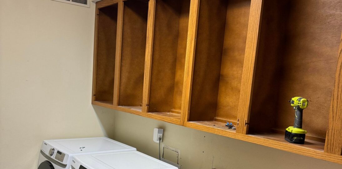 Dated laundry room during demo with honey oak upper cabinets with doors removed, a white Samsung front-load washer and matching dryer side by side, a cordless drill resting on an open shelf, and a bare patch of wall where the hookups will be lowered.