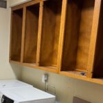 Dated laundry room during demo with honey oak upper cabinets with doors removed, a white Samsung front-load washer and matching dryer side by side, a cordless drill resting on an open shelf, and a bare patch of wall where the hookups will be lowered.