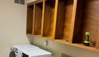 Dated laundry room during demo with honey oak upper cabinets with doors removed, a white Samsung front-load washer and matching dryer side by side, a cordless drill resting on an open shelf, and a bare patch of wall where the hookups will be lowered.