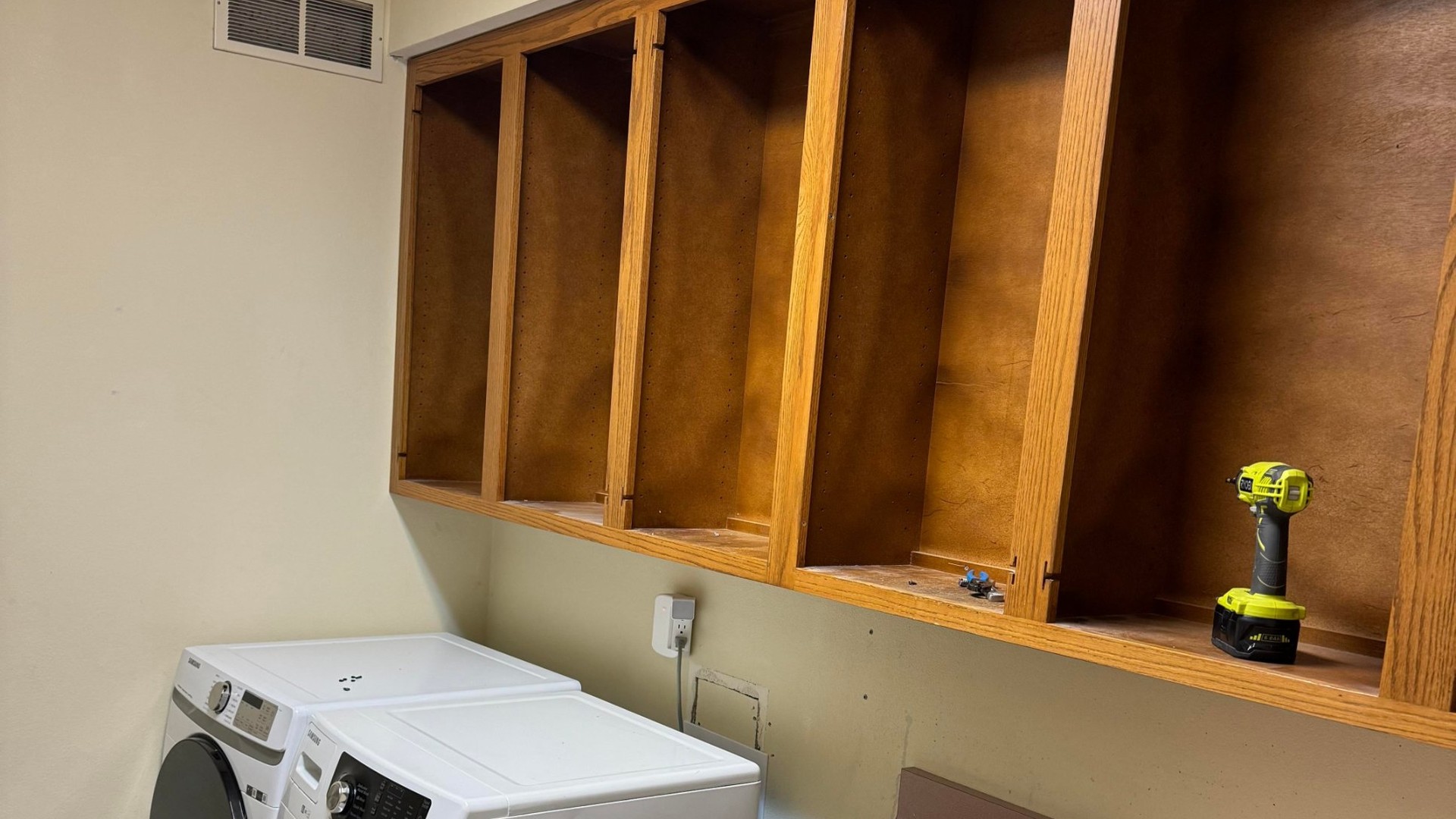Dated laundry room during demo with honey oak upper cabinets with doors removed, a white Samsung front-load washer and matching dryer side by side, a cordless drill resting on an open shelf, and a bare patch of wall where the hookups will be lowered.