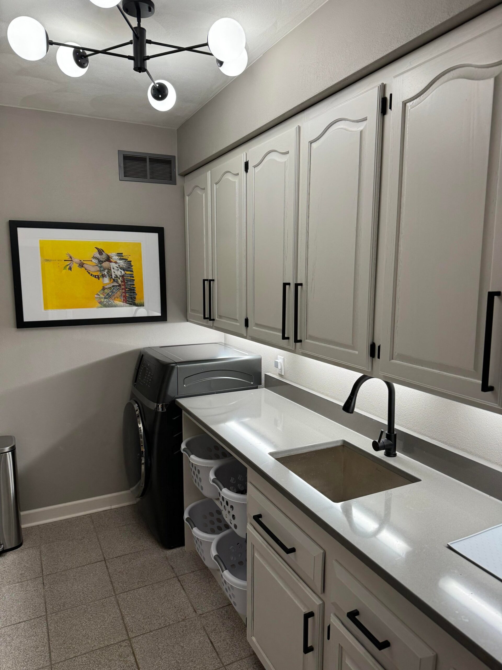 Finished laundry room with light gray cabinets, black bar hardware, gray quartz counter, black undermount faucet over a stainless sink, framed yellow medicine man print on the left wall, and a black sputnik globe light fixture on the ceiling.