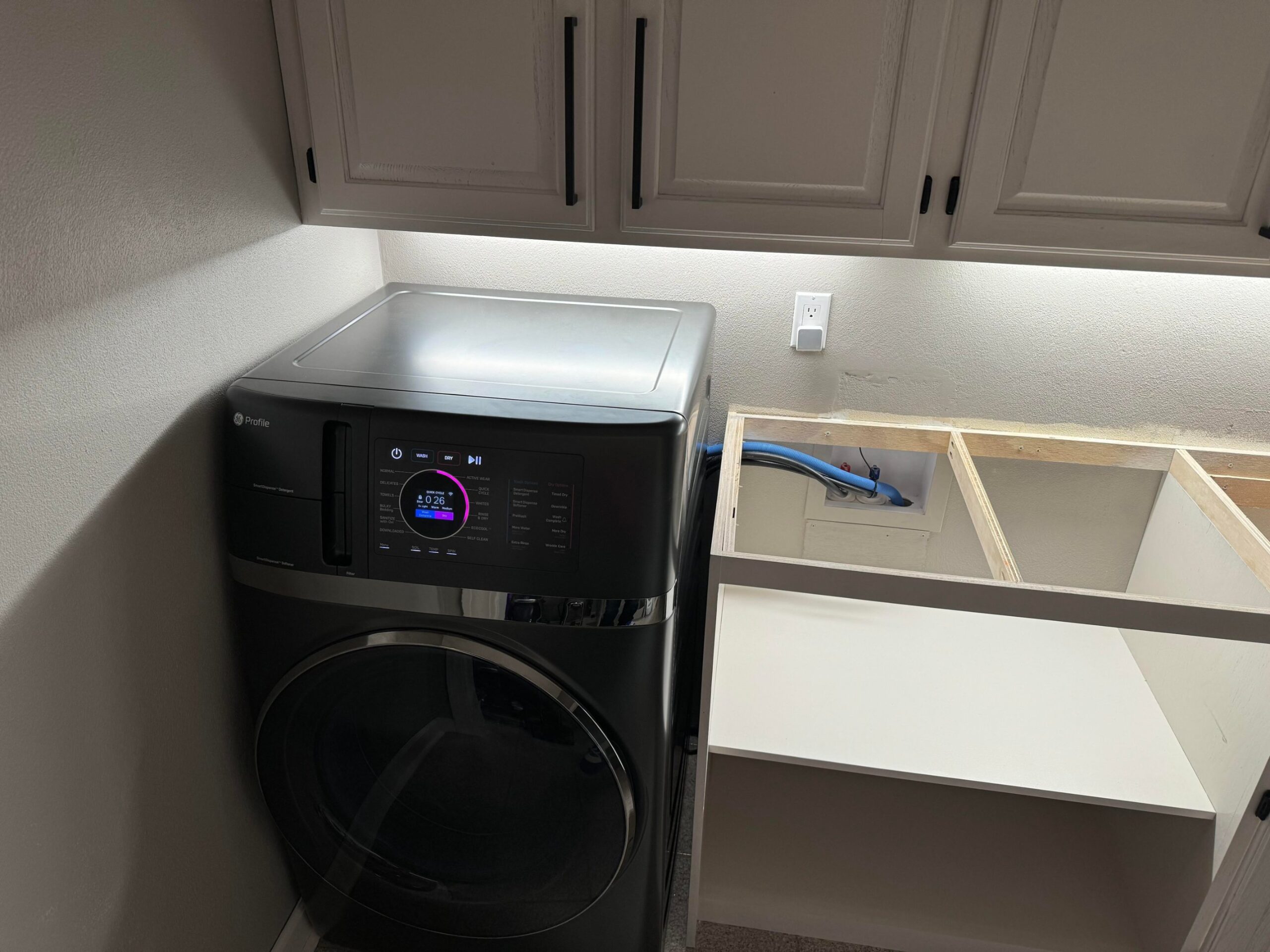 Black GE Profile all-in-one washer dryer combo unit installed in the laundry room, with pink display showing a quick cycle timer, cabinets freshly painted light gray above and the countertop frame open to show plumbing hookups.