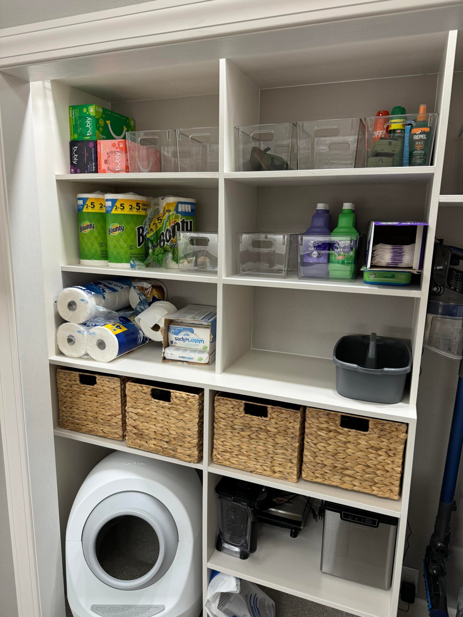 Bottom section of the closet build in with the white Litter Robot self cleaning cat litter box tucked into the lower left cubby next to a Ninja blender and a stainless small appliance, with woven storage baskets above and shelving holding paper towels, toilet paper and cleaning supplies.