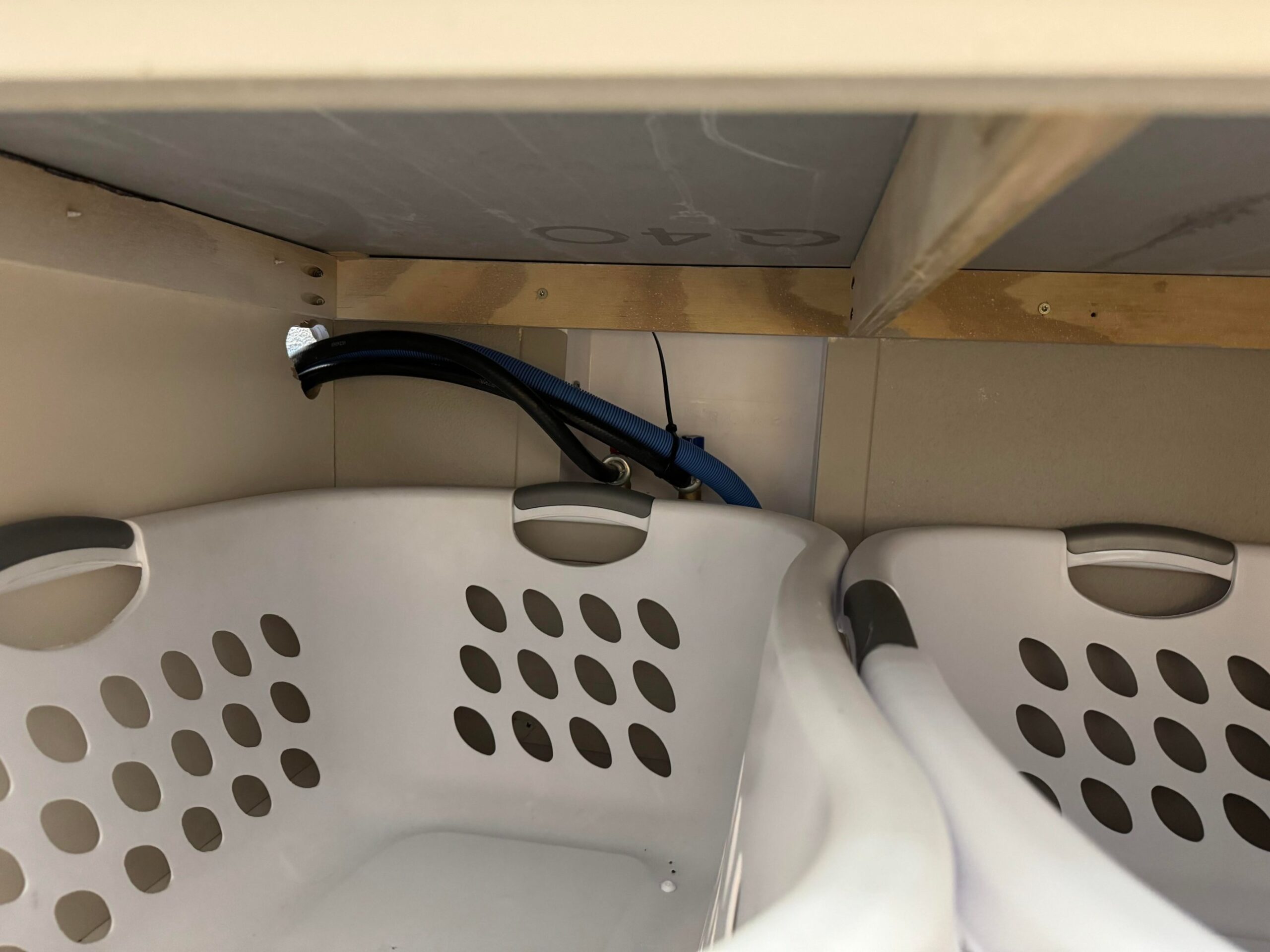 Close up detail of the lowered washer hookups behind the laundry basket cubbies showing a blue hot water flex hose, braided cold line and black drain hose zip tied together at a low height inside the plywood framing.
