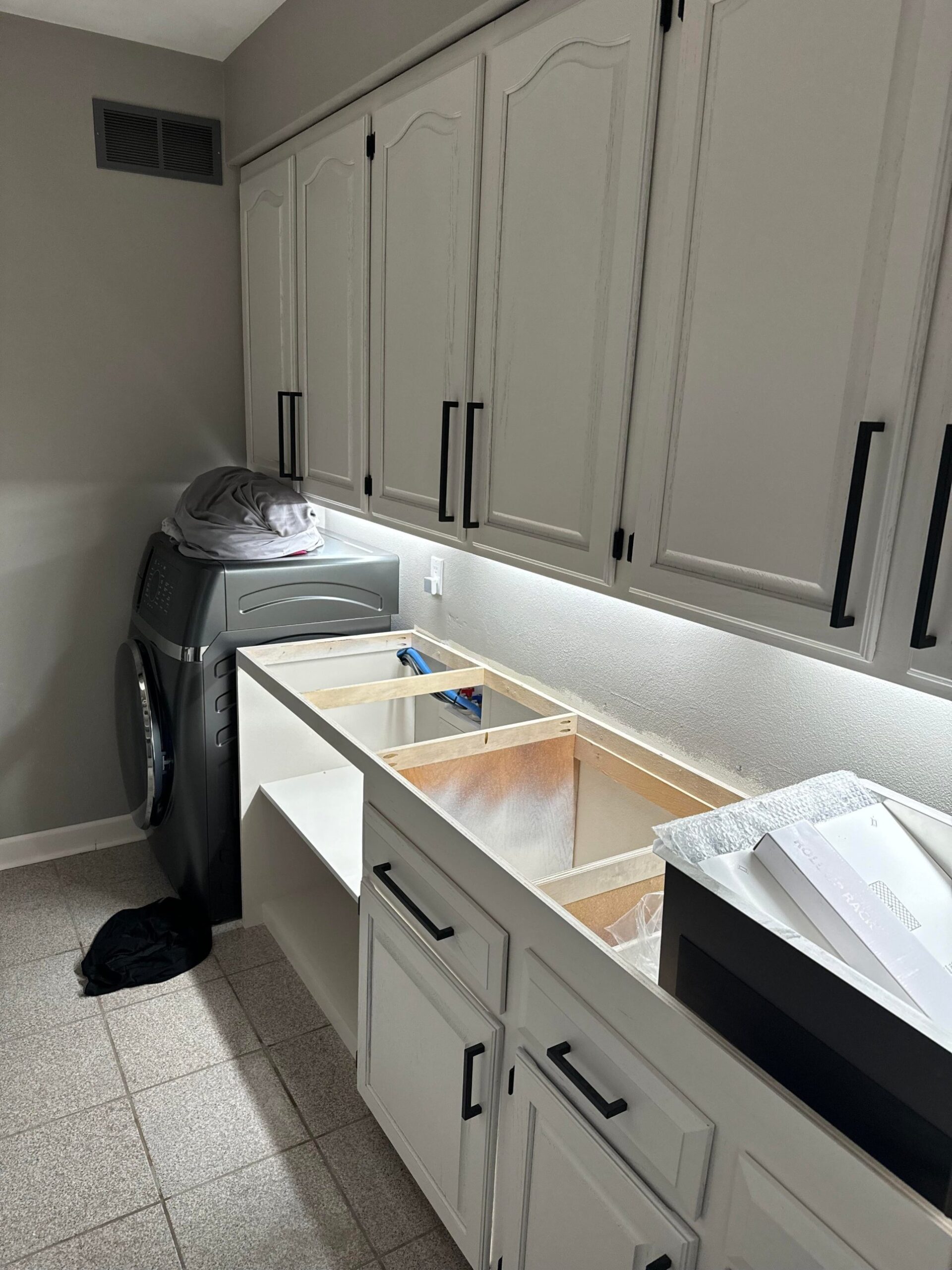Laundry room mid-remodel with cabinets freshly painted light gray, black bar pulls installed, old washer still in place, and a demo cavity cut into the counter where a new sink and basket cubby will go.