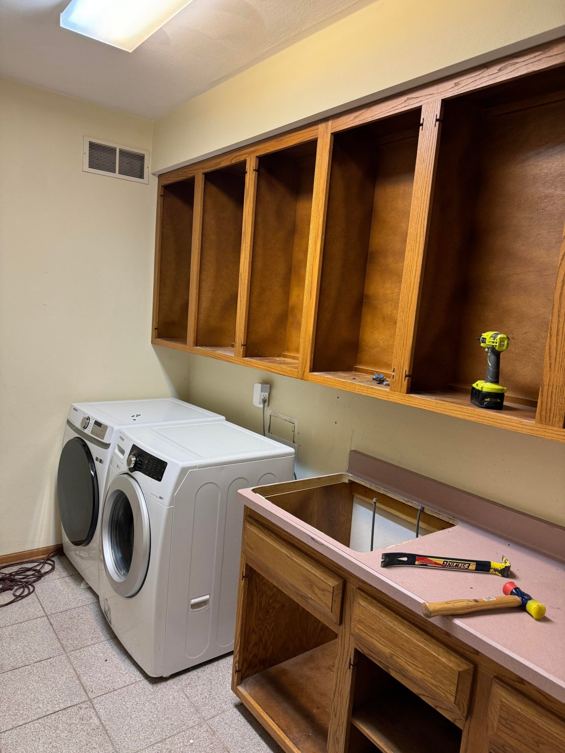 Original laundry room with pink laminate counter top ripped open, honey oak upper cabinets with doors off, white Samsung front-load washer and matching dryer, drill resting on a shelf, hammer and pry bar on the counter.