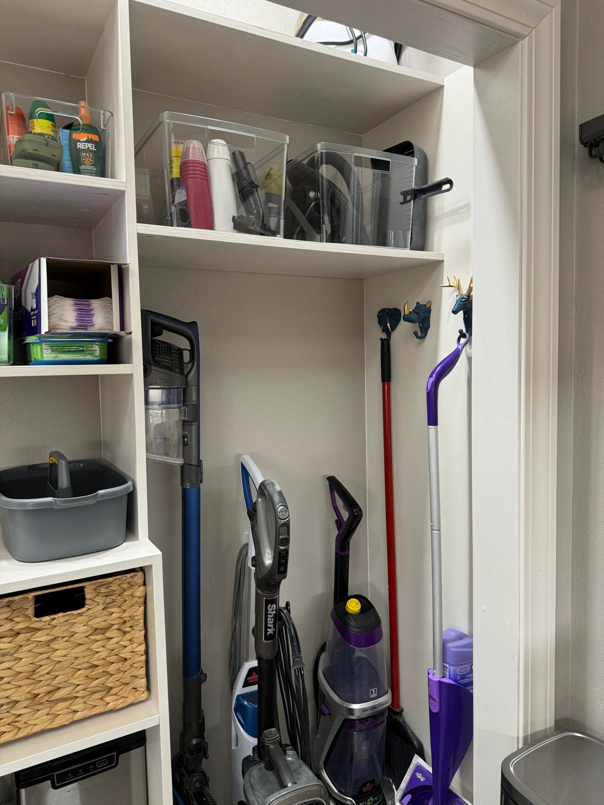 Close up of the right side of the built in closet showing a Shark stick vacuum, a Shark upright, a Bissell Pet Pro carpet cleaner, a broom and a Swiffer wet jet all standing side by side against the wall, with navy and gold animal head wall hooks holding the broom and Swiffer handles upright, and clear bins of insect repellent and travel cups on the top shelf.