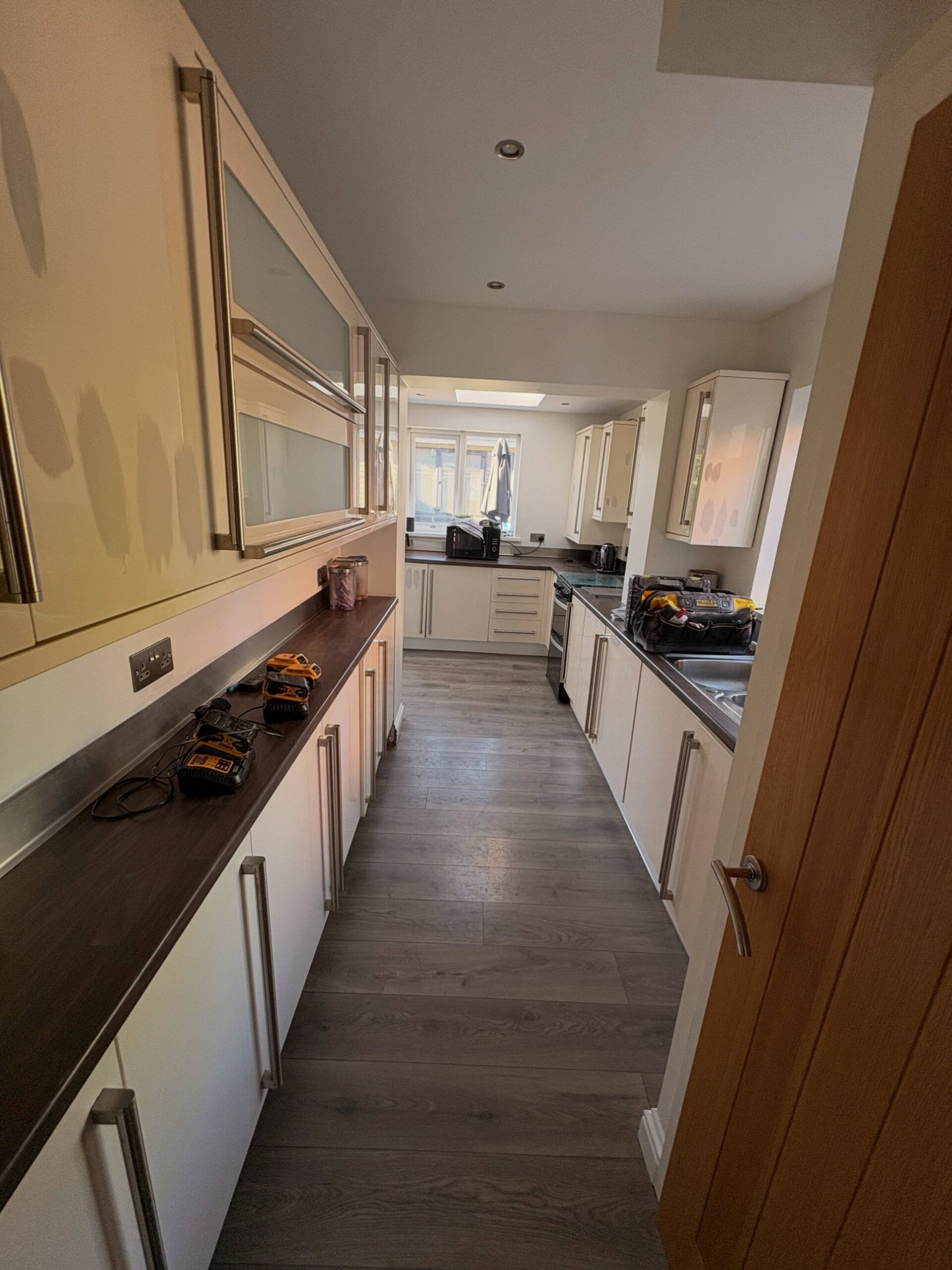 Long galley kitchen with cream cabinets showing patchy marks and shadows on the upper doors, dark walnut counters, long vertical brushed handles, a tool bag and battery chargers set out on the counter.
