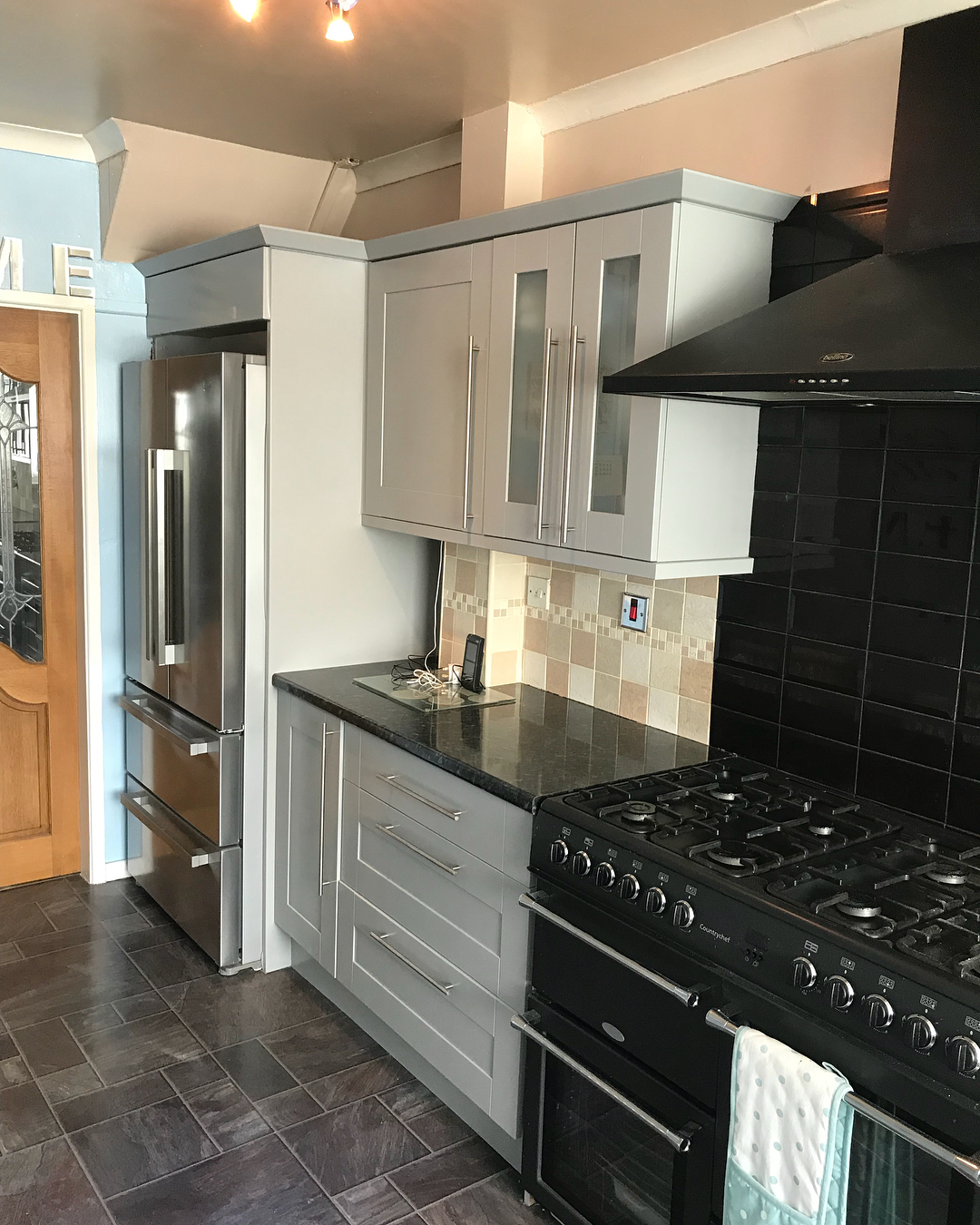 The painted gray kitchen showing the tall fridge freezer, upper cabinets with frosted glass doors, and the same black range cooker, now set against a cool, cohesive color story.