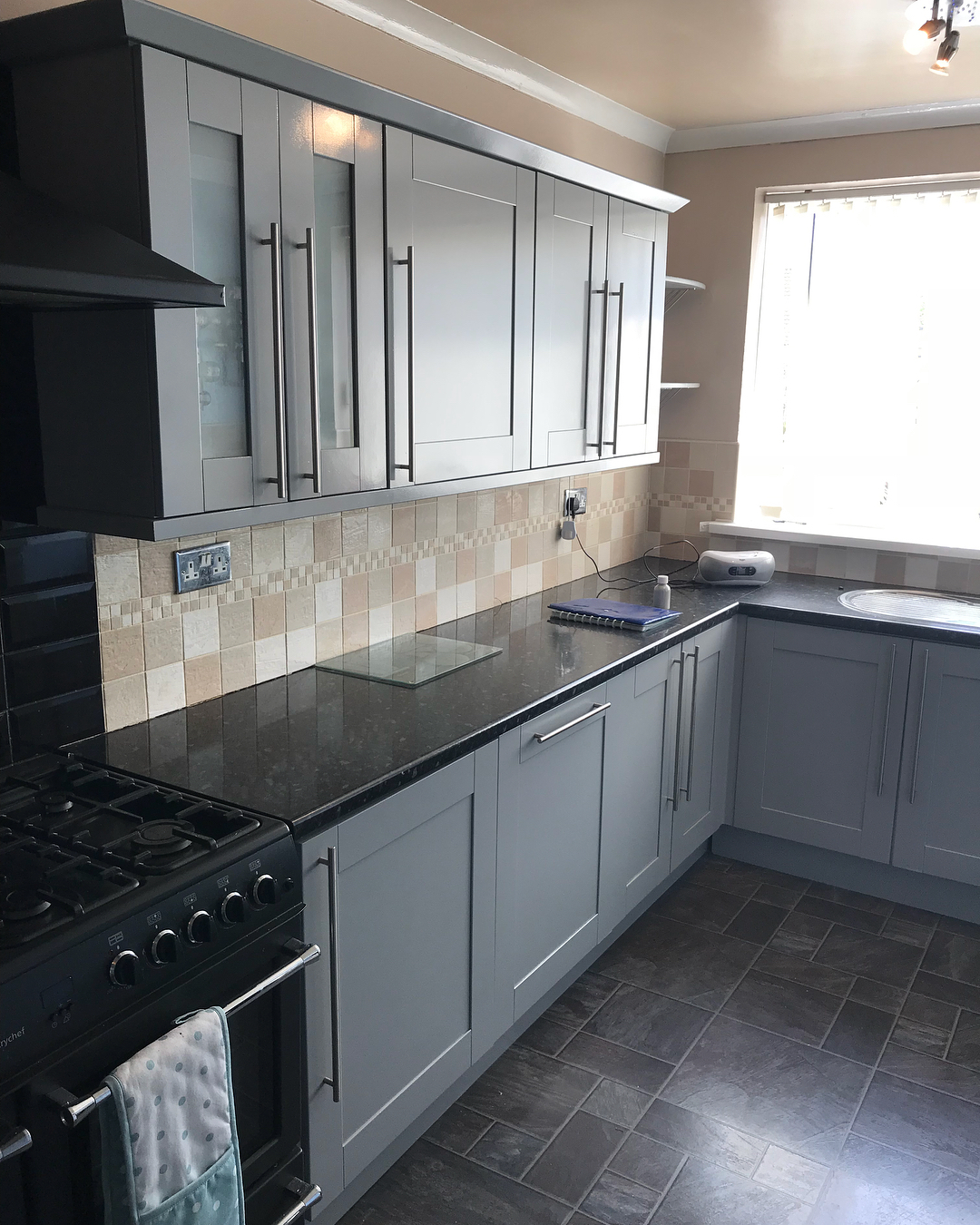 Finished gray painted L-shaped kitchen with the same mosaic backsplash, black granite counters, slate floor, and bright window on the right, now looking current and bright.