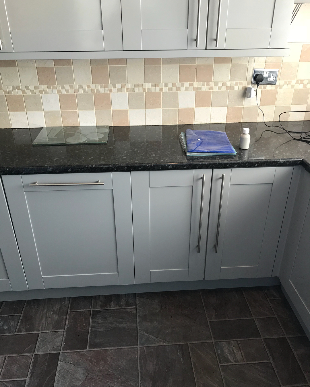 Lower gray painted Shaker cabinets with slim brushed steel handles, dark granite counter, and the slate style tile floor stretching across the room.
