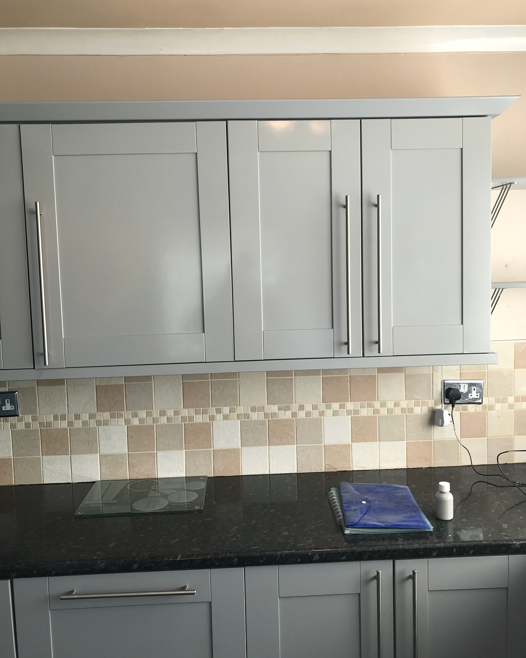 Three Shaker upper cabinets in Manor House Grey with brushed steel bar handles, above the warm cream and beige mosaic tile backsplash and dark granite counter.