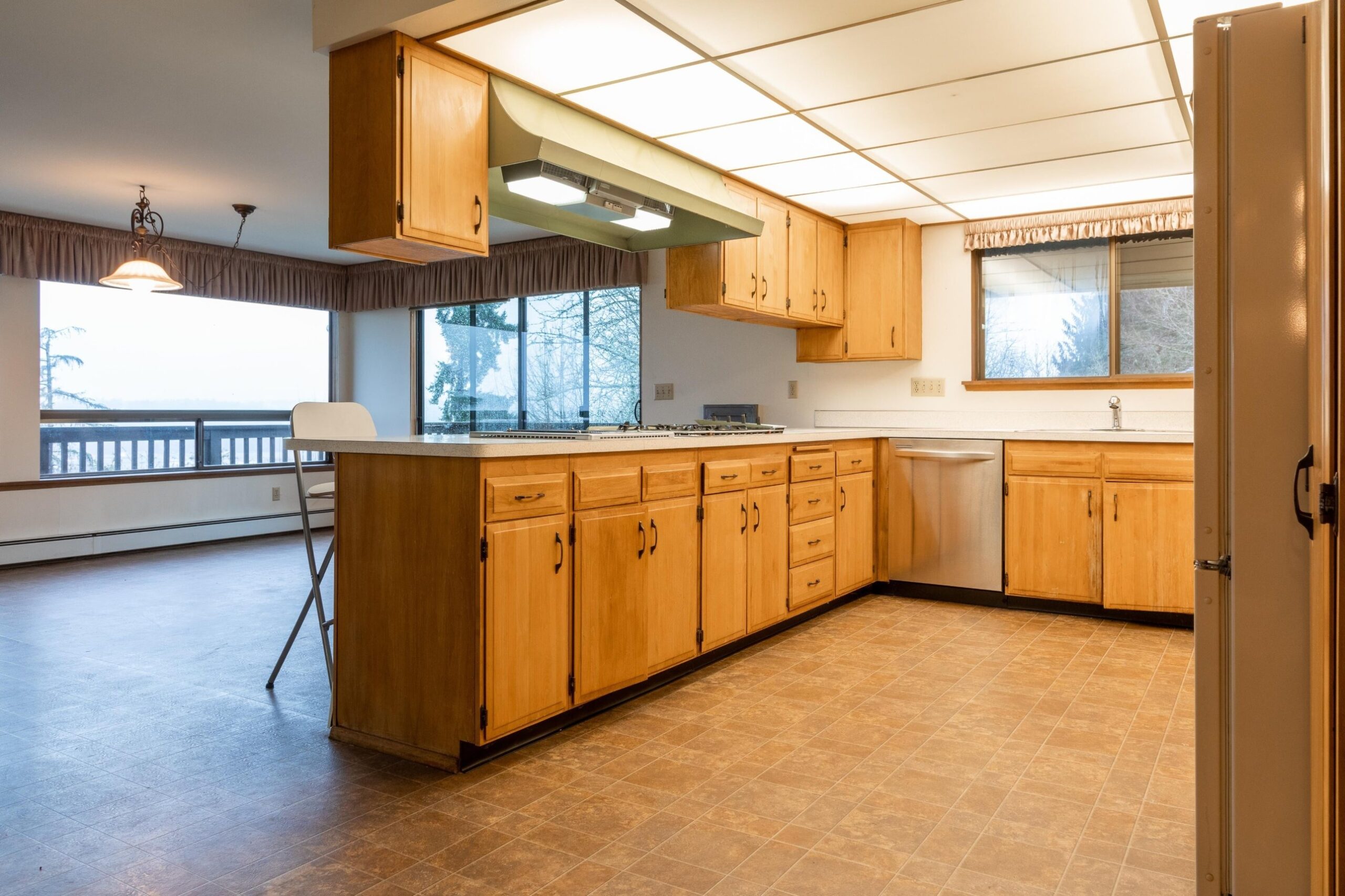 Wide view of a dated kitchen with light maple cabinets, a dropped ceiling soffit holding a fluorescent light panel, pleated fabric valances over the windows, tan tile floor, and a dining area to the left.