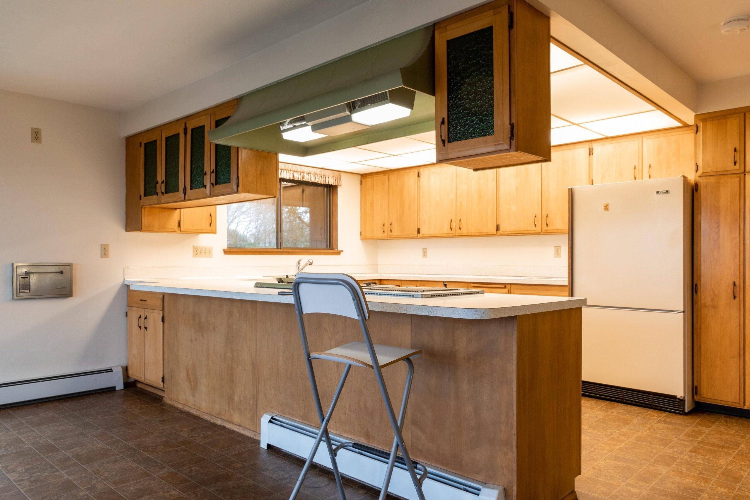View of the same kitchen's peninsula from the opposite angle, showing a sage green range hood, white fridge, green-tinted glass cabinet inserts, a pleated valance over a small window, and a folding bar stool at the counter.