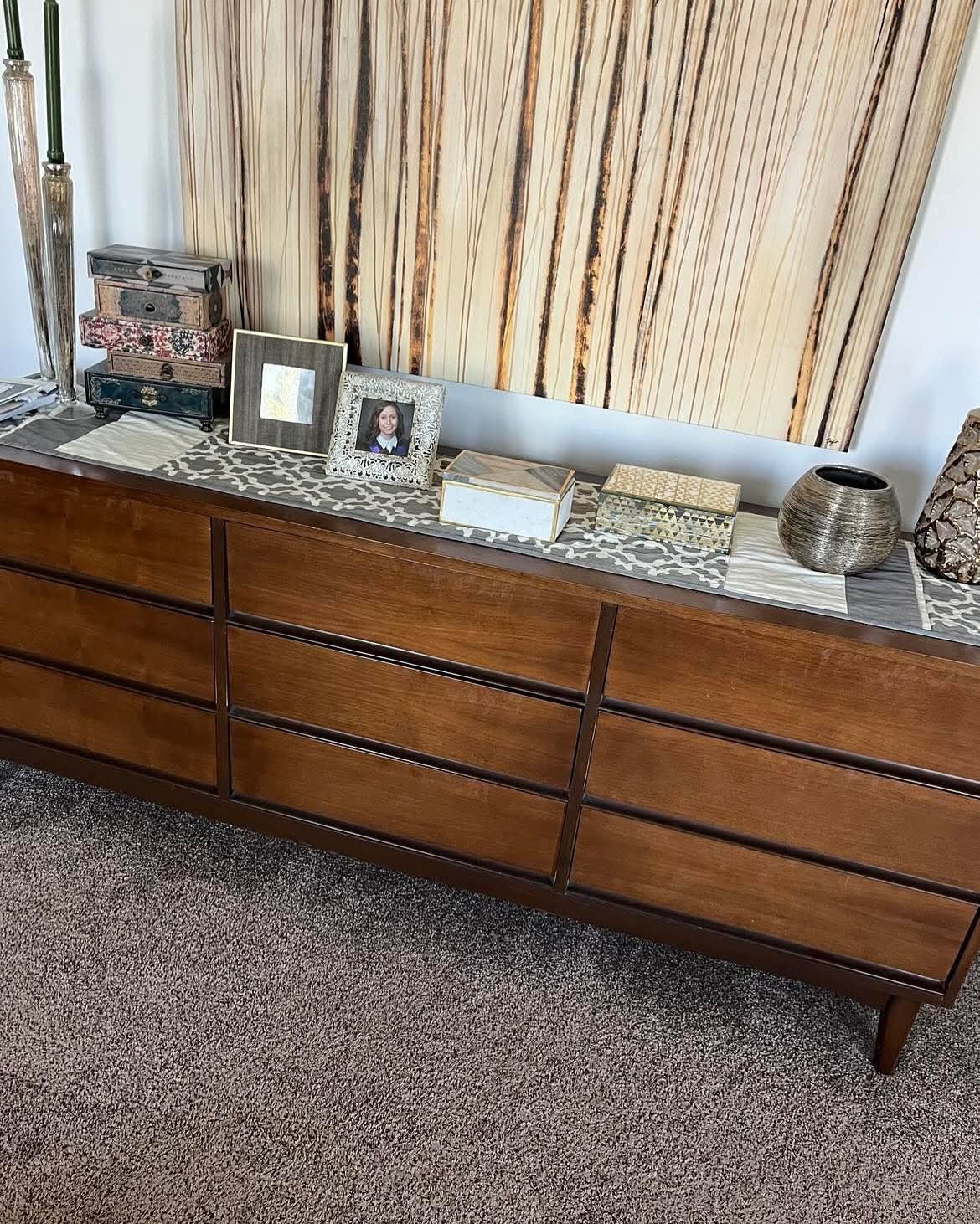 Long nine-drawer mid-century modern walnut dresser in dark stain, styled with jewelry boxes, picture frames, and a metallic vase against a textured wall hanging, on brown carpet.