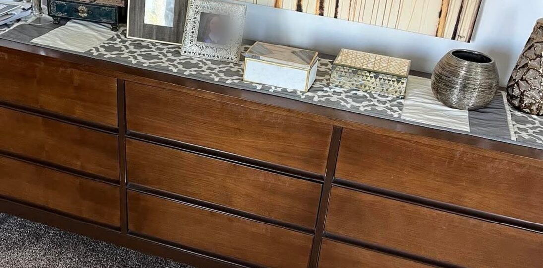 Long nine-drawer mid-century modern walnut dresser in a dark orange-brown stain, styled with jewelry boxes, picture frames, and a metallic vase along the top, shown before refinishing in a 1974 solid walnut bedroom set.
